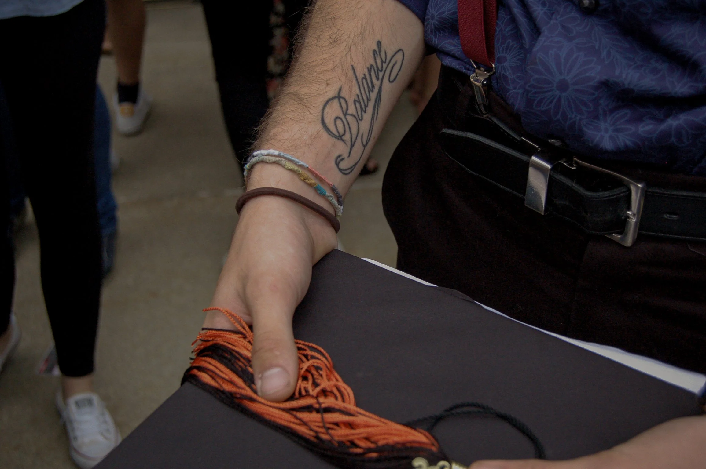A tattoo on a person's arm reading 'Benjamin' in script. The person is wearing a blue shirt with a pattern, black pants, and has bracelets on their wrist. They are holding hands with another person, who has orange and black shoelaces visible.