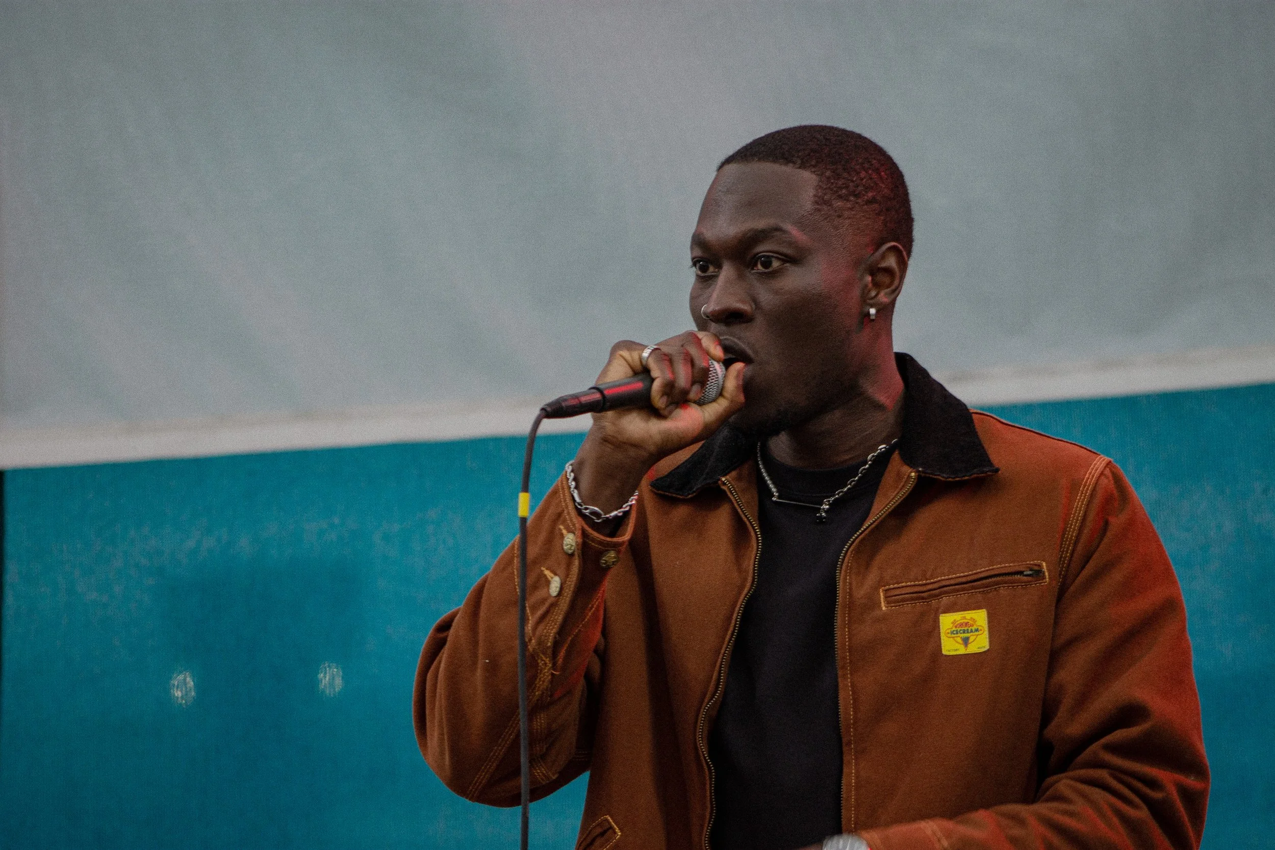 A young man with dark skin and short hair holding a microphone, wearing a brown jacket with a yellow patch, a silver chain, and earrings, standing in front of a teal and gray wall.
