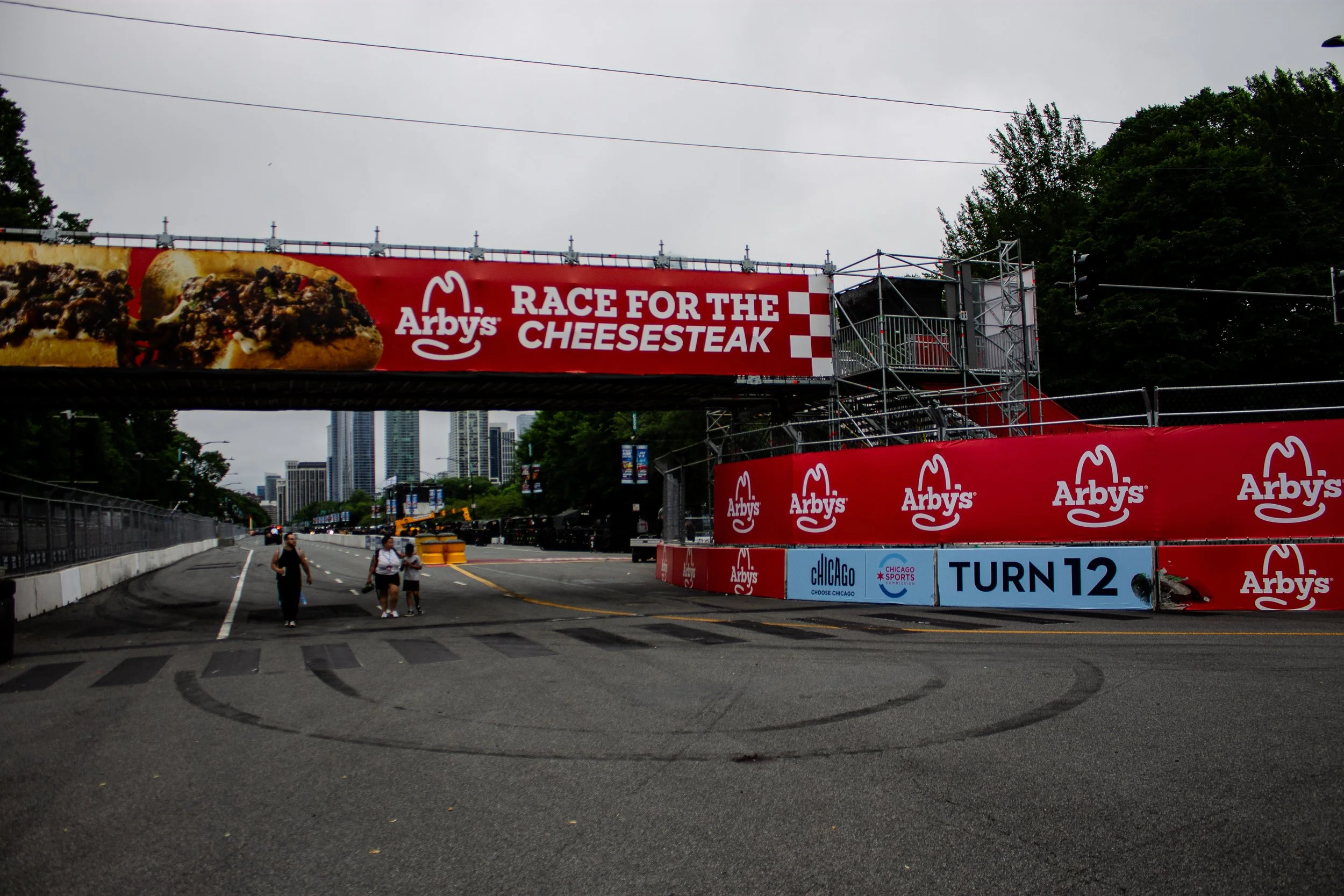 Street view of an outdoor event area with a large red banner advertising Arby's 'Race for the Cheesesteak' race, featuring a cheesesteak sandwich image. The area is set up for a race with a start/finish line, and a few people walking nearby against a backdrop of skyscrapers and trees on a cloudy day.
