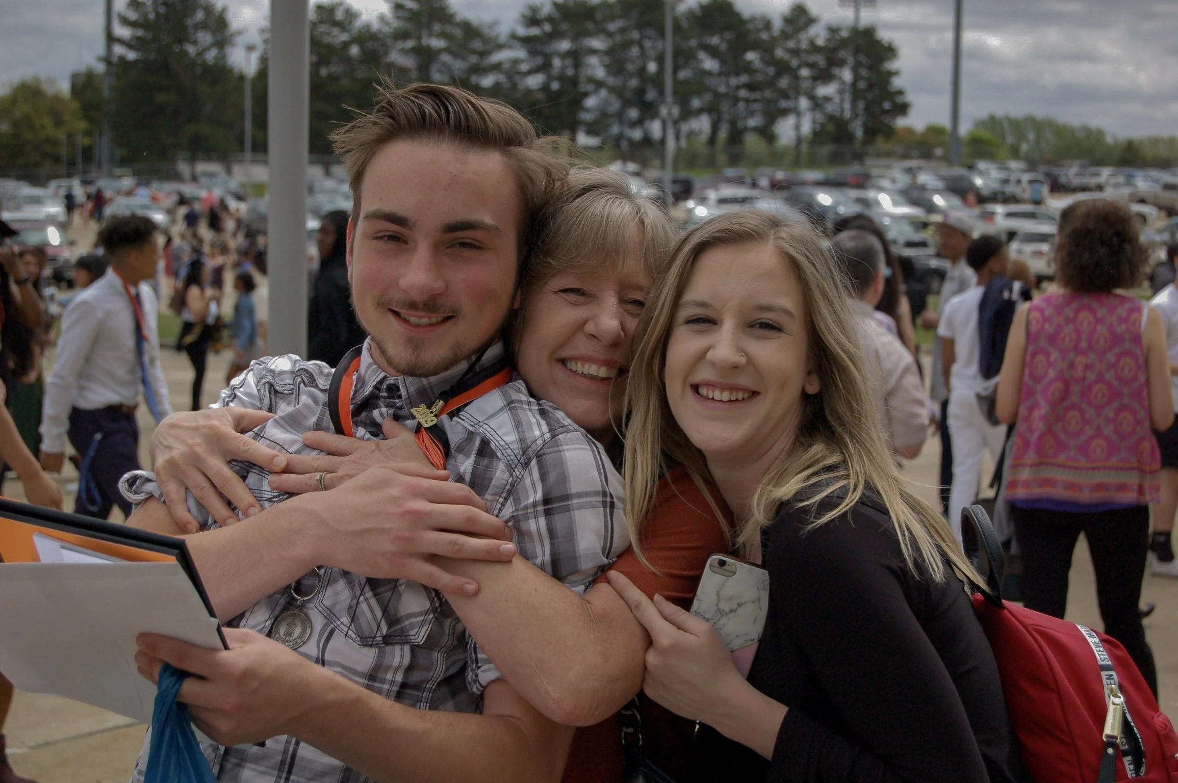 Three people hugging each other and smiling at the camera at an outdoor event with many people and parked cars in the background.