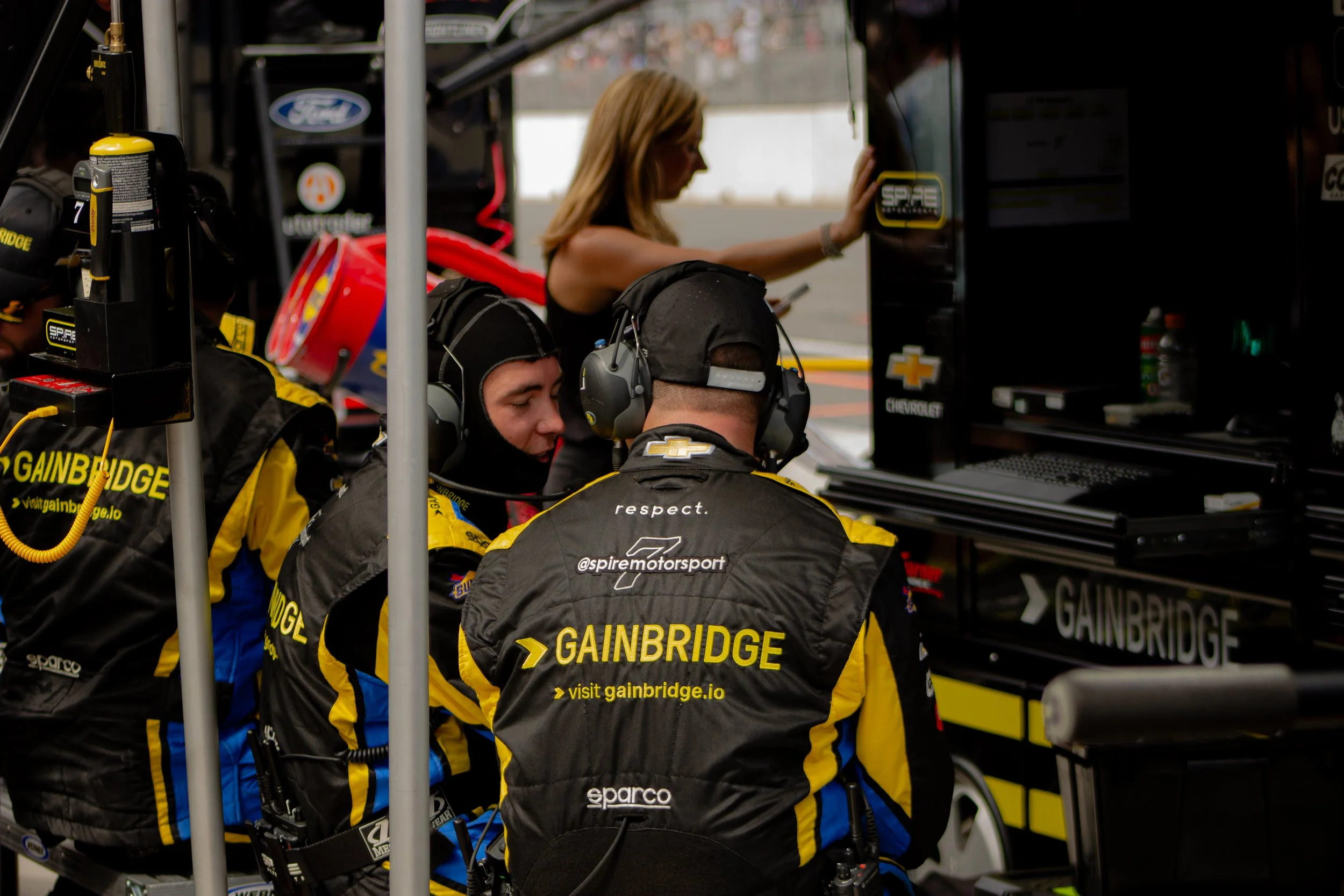 Race car team members in yellow and black uniforms working in the garage, with racing equipment and a woman using a smartphone in the background.