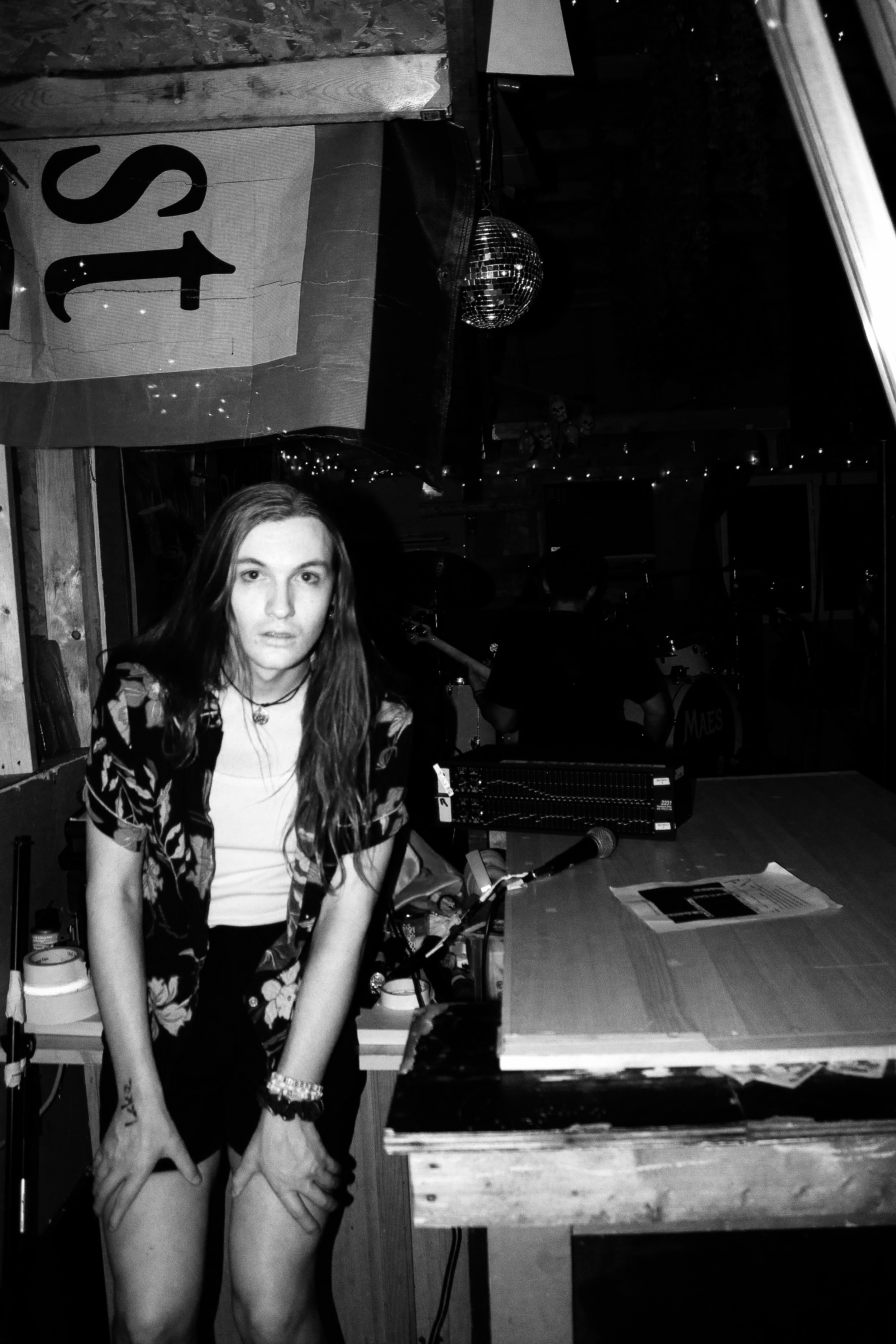 A young woman with long hair wearing a floral shirt, sitting at a table in a dimly lit room with a disco ball hanging from the ceiling, a sign with the number 45, and other people in the background.