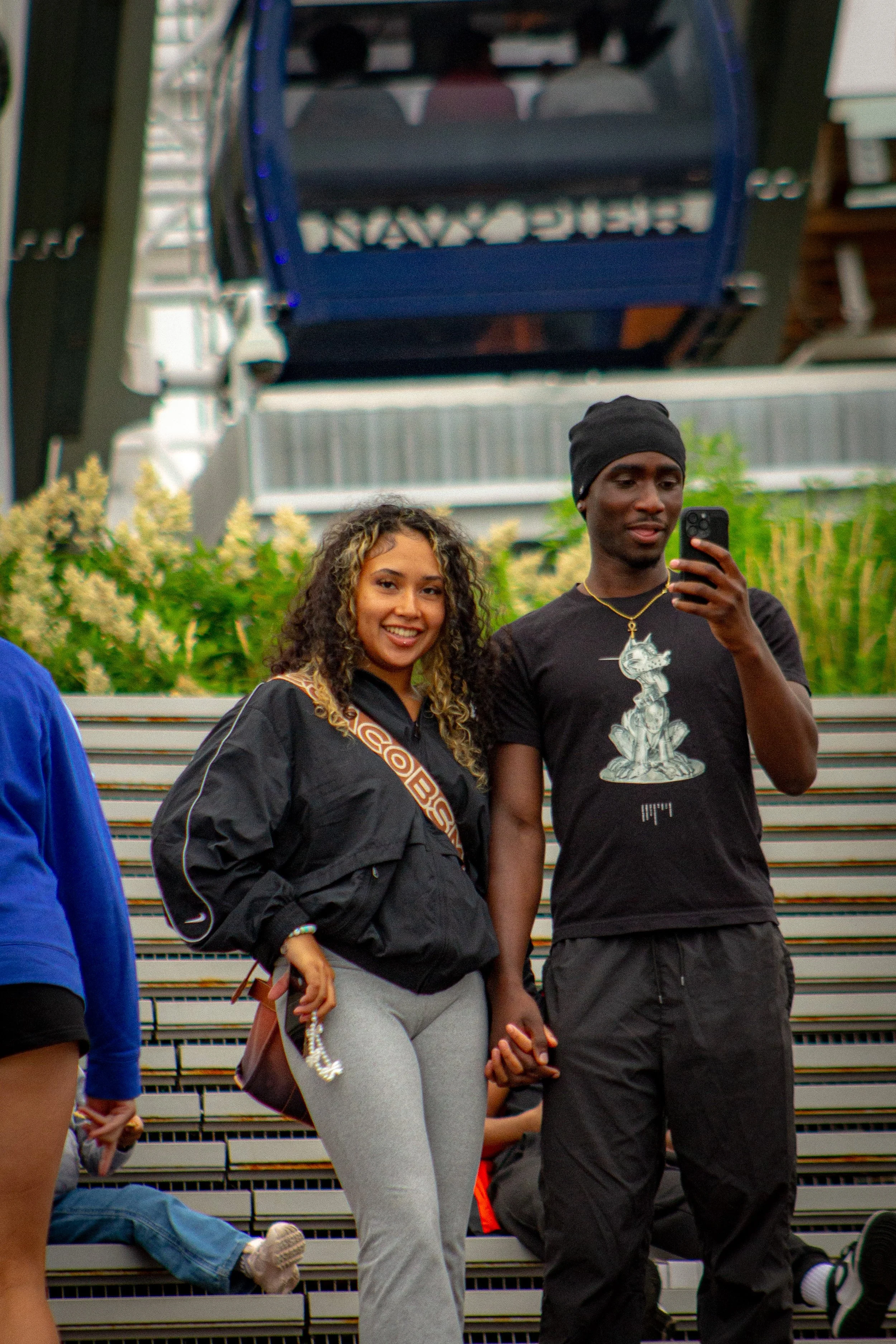 Two young adults, a woman with curly hair and a man wearing a black beanie, pose for a selfie on outdoor bleachers with others seated behind them and green foliage in the background.