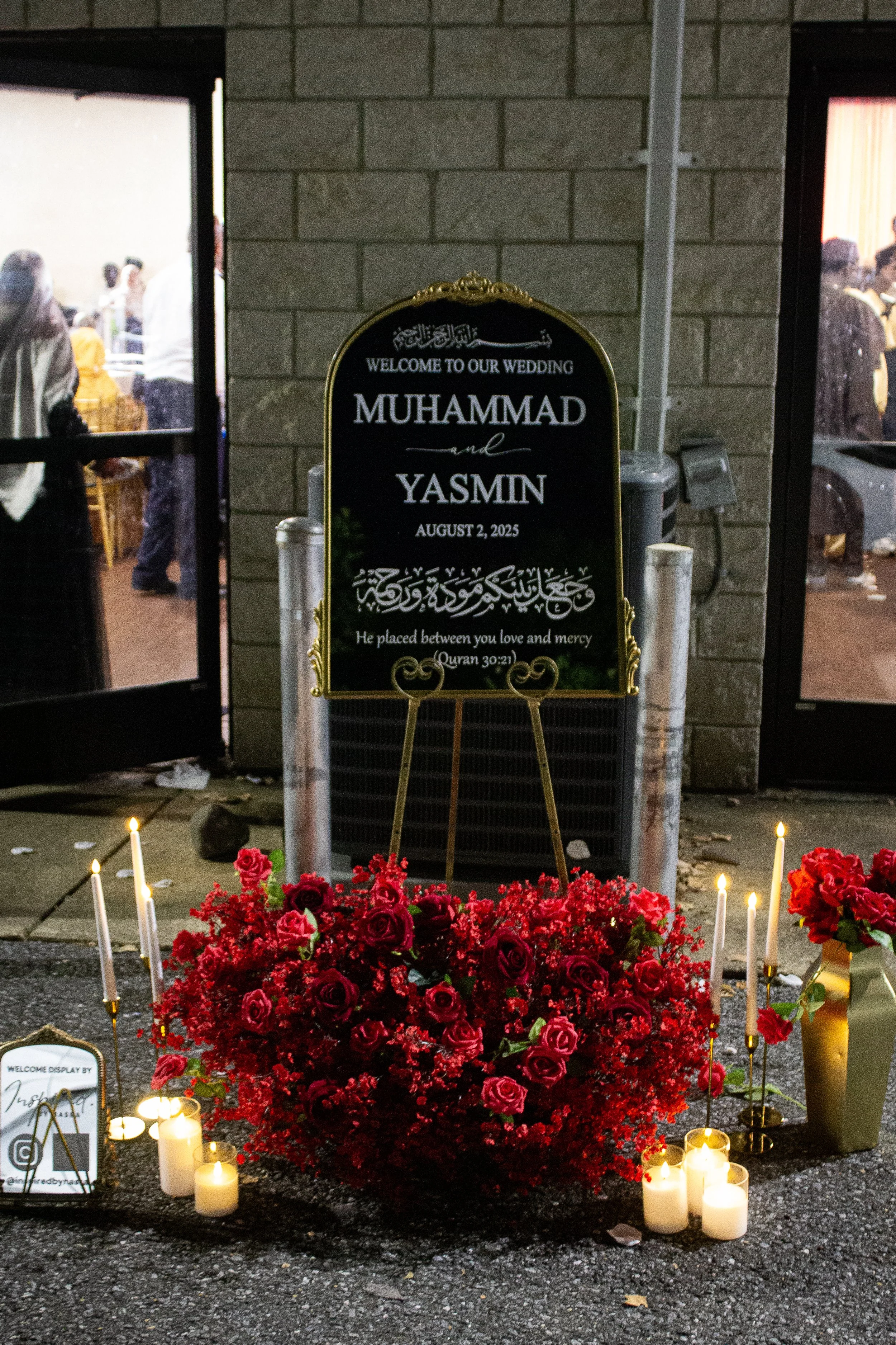 Flowers, candles, and a memorial board at a wedding mourning site for Muhammad and Yasmin on August 2, 2025, with people visible through the windows.