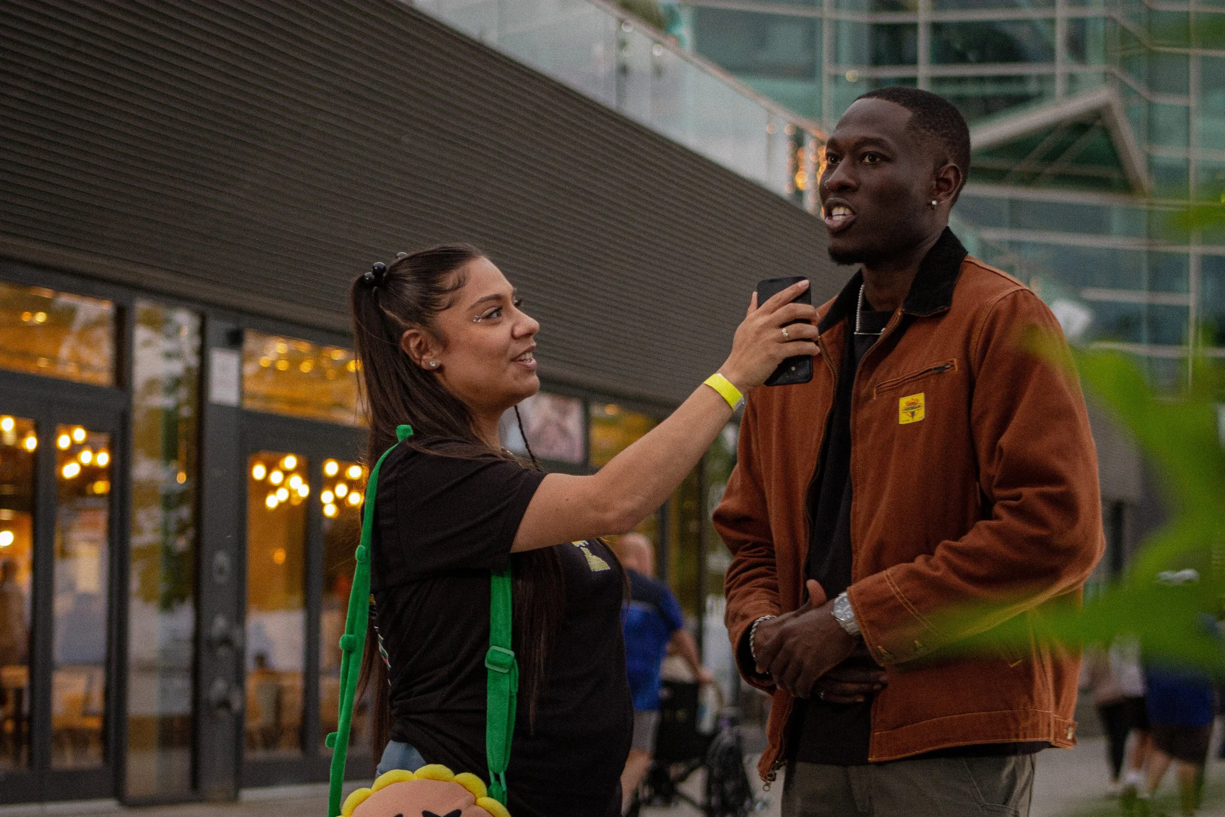A woman with long dark hair, wearing a black t-shirt and green apron, holds a microphone and explains something to a tall man with short dark hair, wearing a brown jacket, in an outdoor setting near a modern building with glass windows and lights.