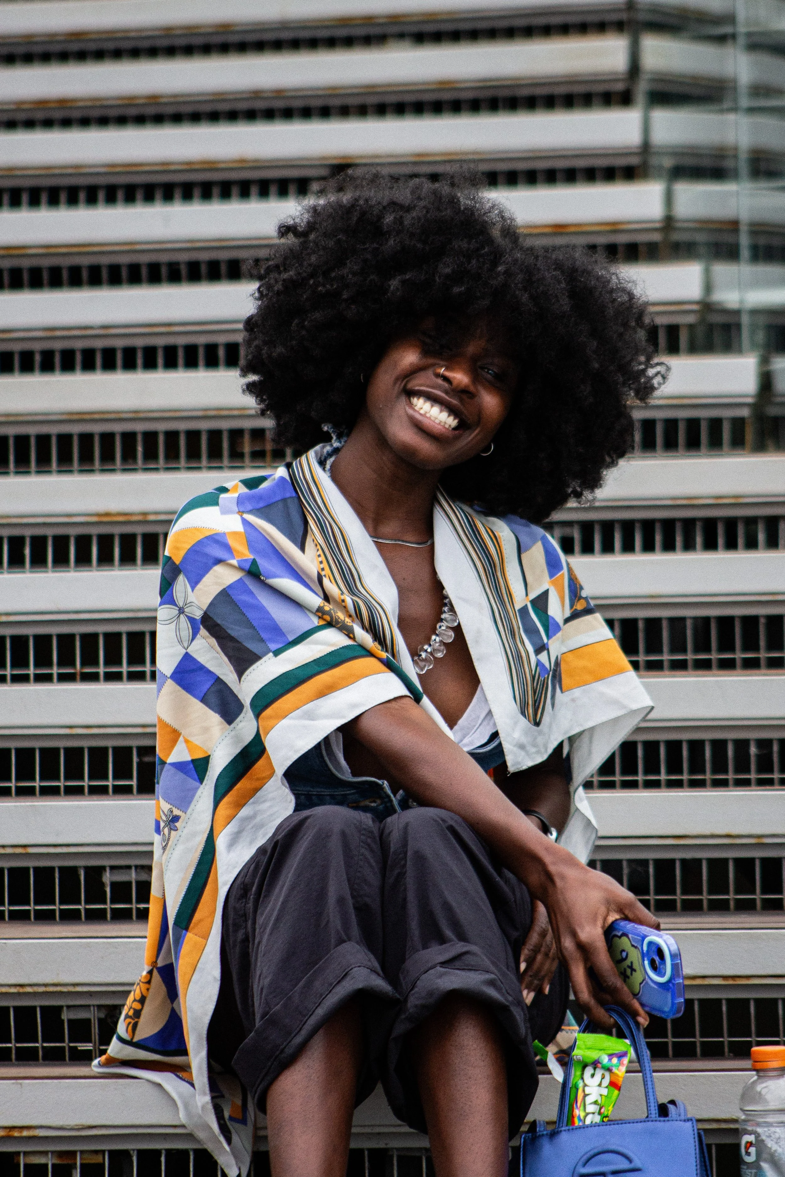A woman with a large curly afro hairstyle is sitting on outdoor bleachers, smiling at the camera. She is wearing a colorful, patterned button-down shirt and black shorts. She has a nose ring and is holding a blue tablet, with snacks and drinks in nearby bags.