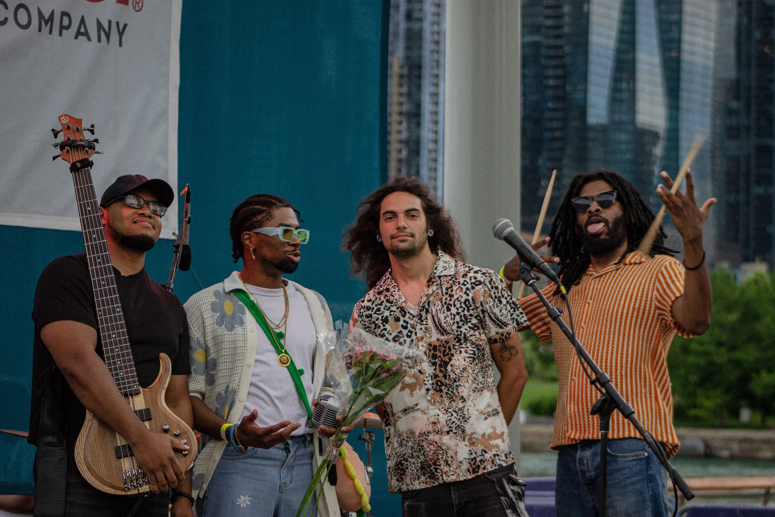 Four men standing on a stage outdoors, one holding a guitar, another holding a microphone, one with a bouquet of flowers, and another making a gesture with his hand. They are in front of an urban backdrop with tall buildings.