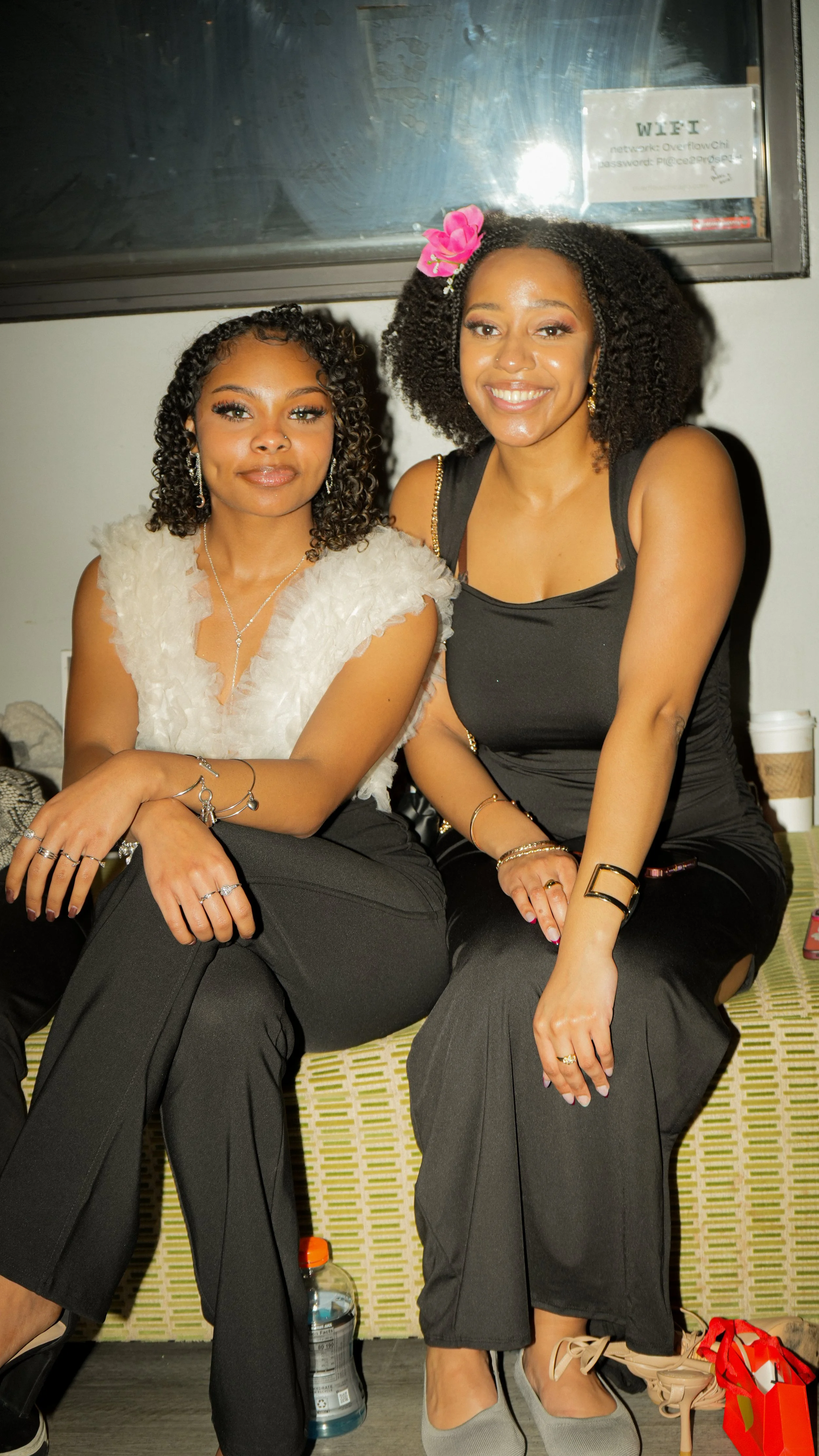 Two women sitting together and smiling at the camera inside a room. They are dressed casually with jewelry and have curly hair. A pink flower accessory is on the woman on the right. A water bottle is on the floor beside their feet.
