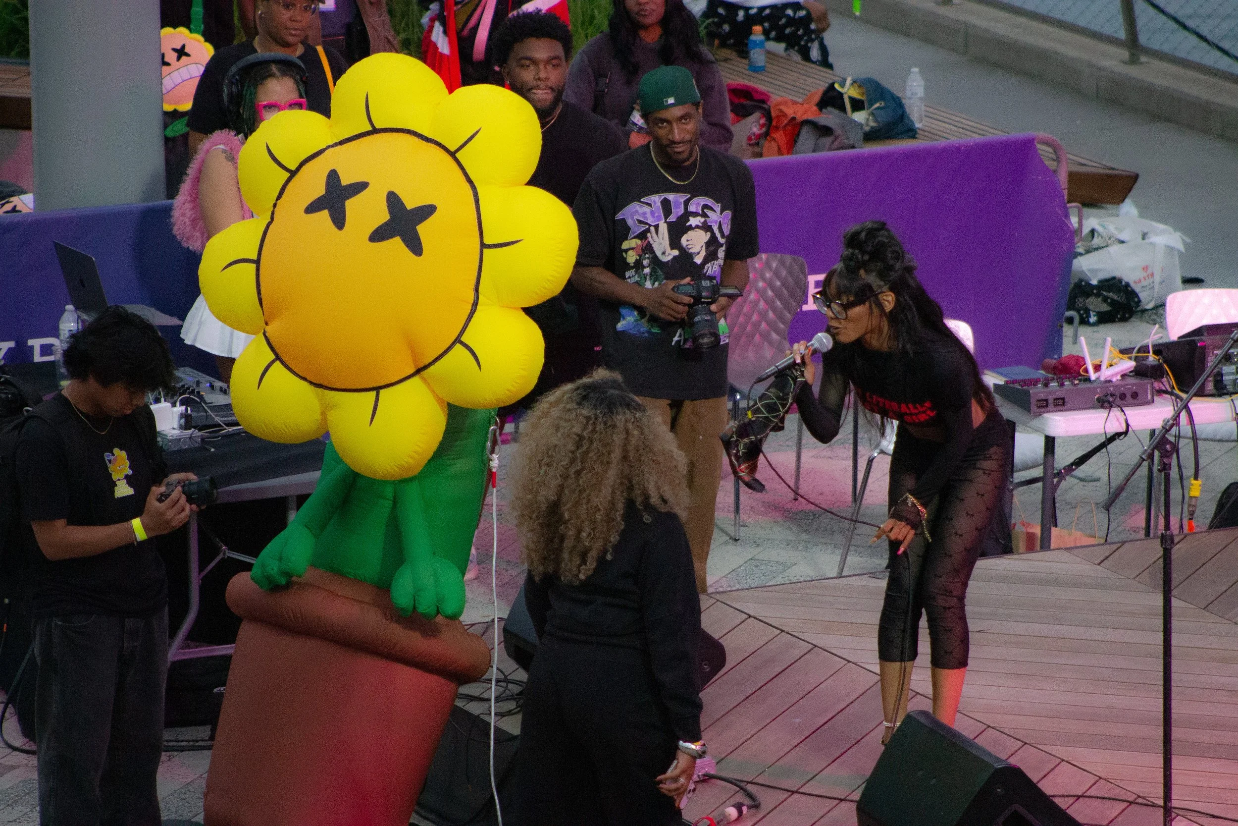 People gathered at an outdoor event, with a large inflatable yellow flower with a face as the centerpiece. A woman is singing into a microphone, and a man is holding a camera. Various people are visible in the background, some seated and some standing, with drinks and equipment around.