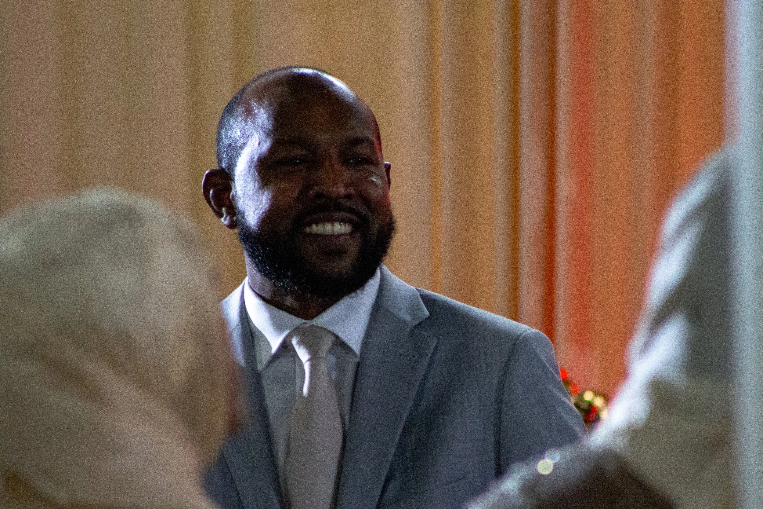 A smiling man in a gray suit and white shirt at a party or social gathering, with blurred decorations in the background.