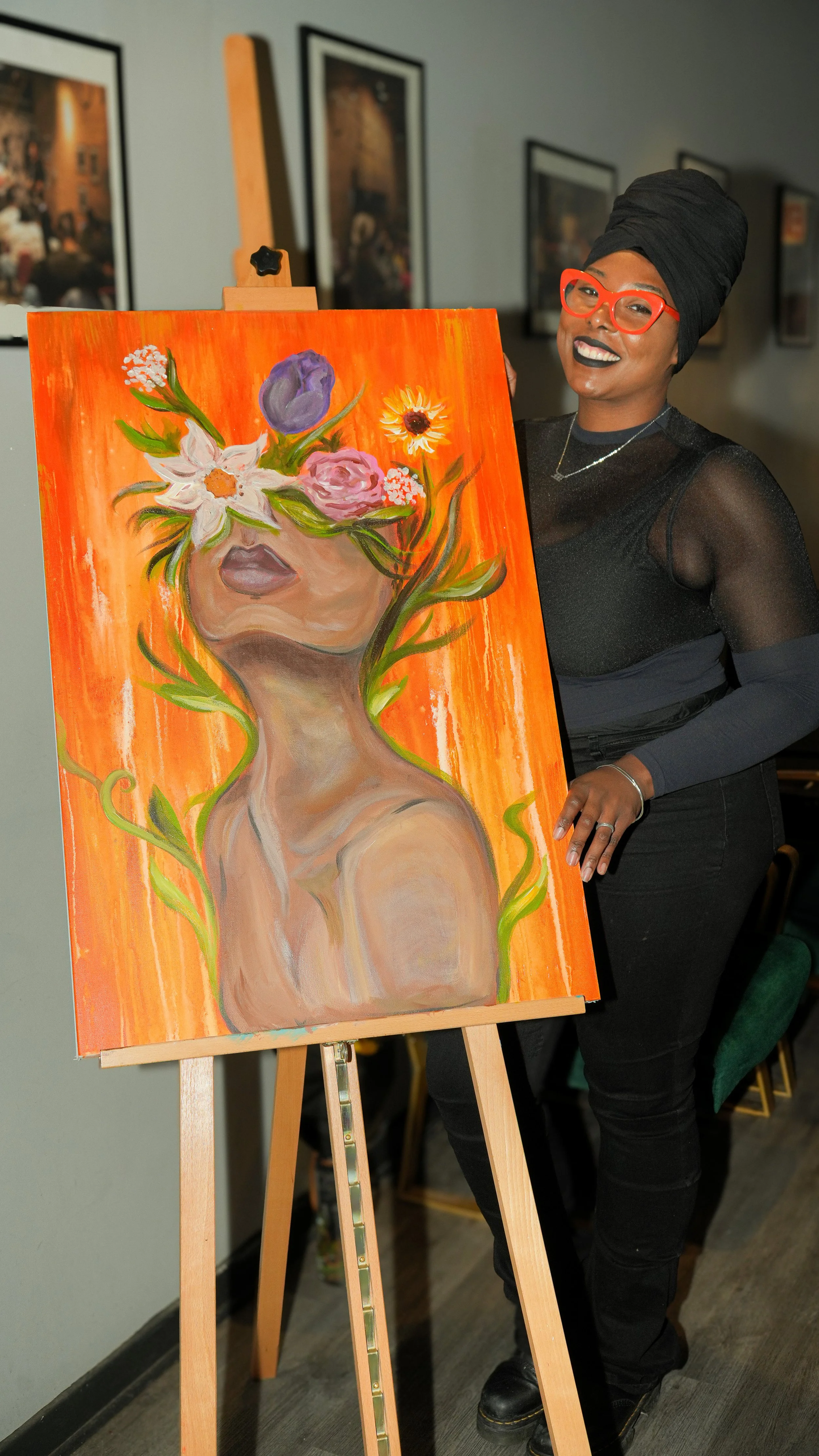 A woman with a black headwrap, red glasses, and a black sheer top is standing next to an easel with a colorful painting of a woman's face covered with flowers. The background features framed pictures on the wall.