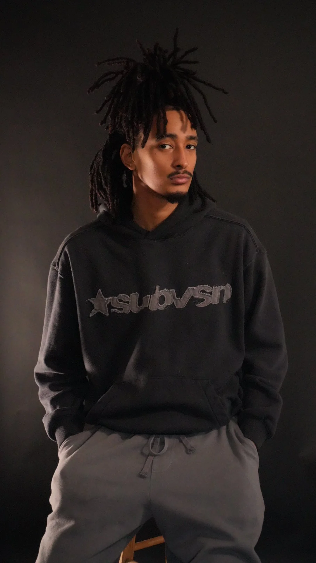 A young man with dreadlocks wearing a black hoodie and gray sweatpants against a plain dark background.