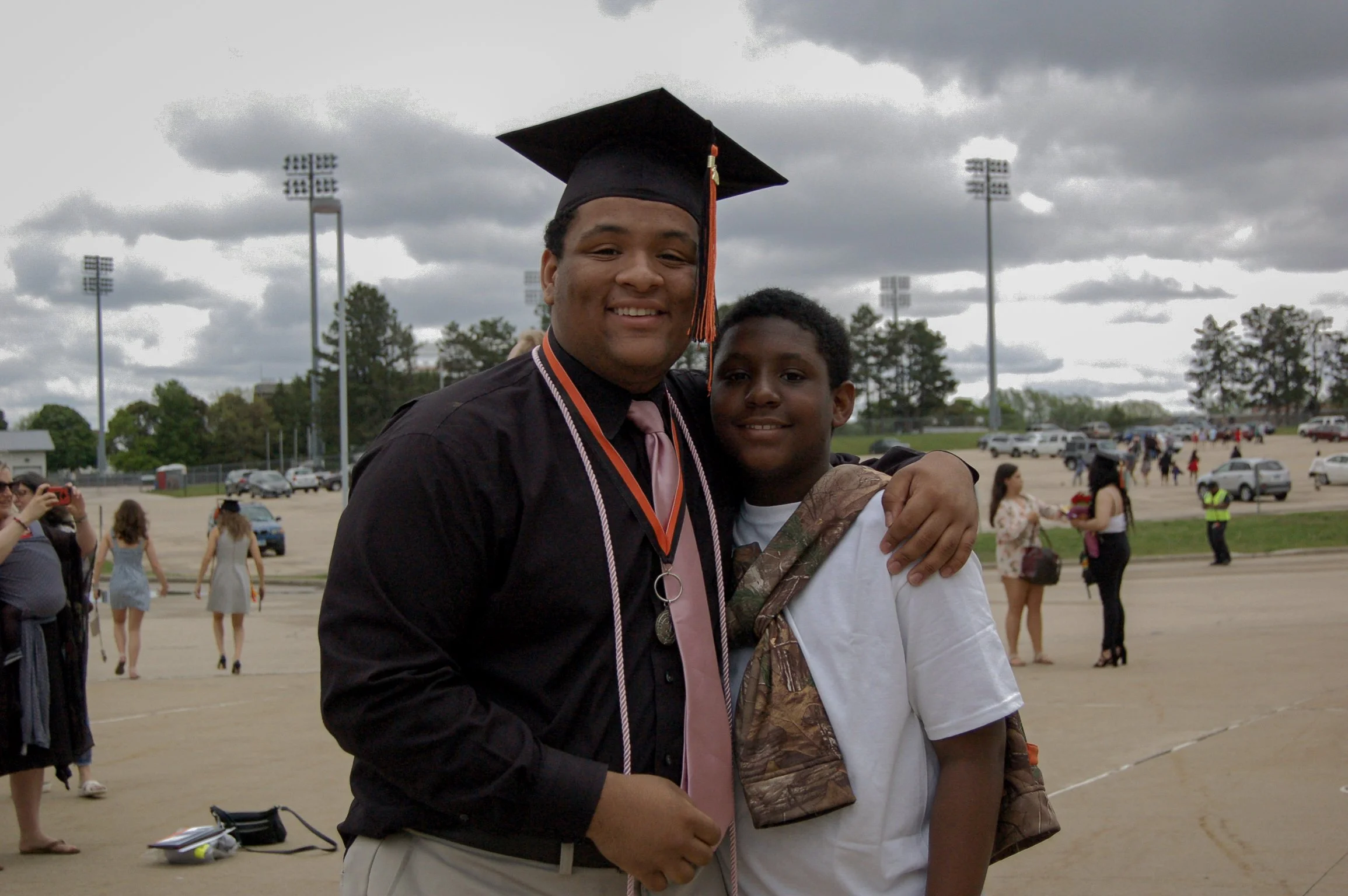 A young man in a graduation cap and gown standing next to a boy, both smiling, at an outdoor graduation ceremony on a cloudy day.