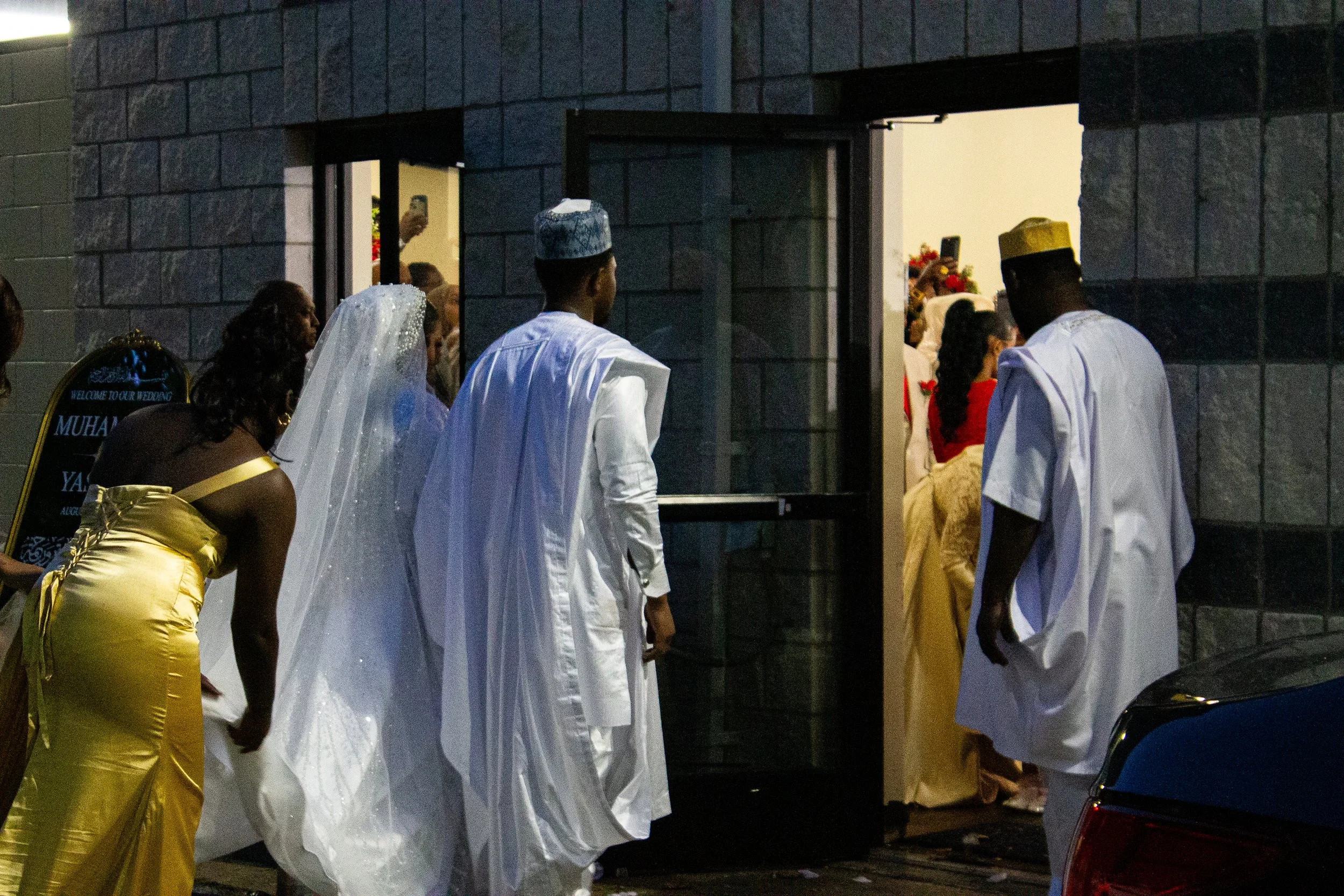 People dressed in traditional Nigerian attire at an indoor event, possibly a wedding or celebration, with some wearing white and gold garments and headpieces.