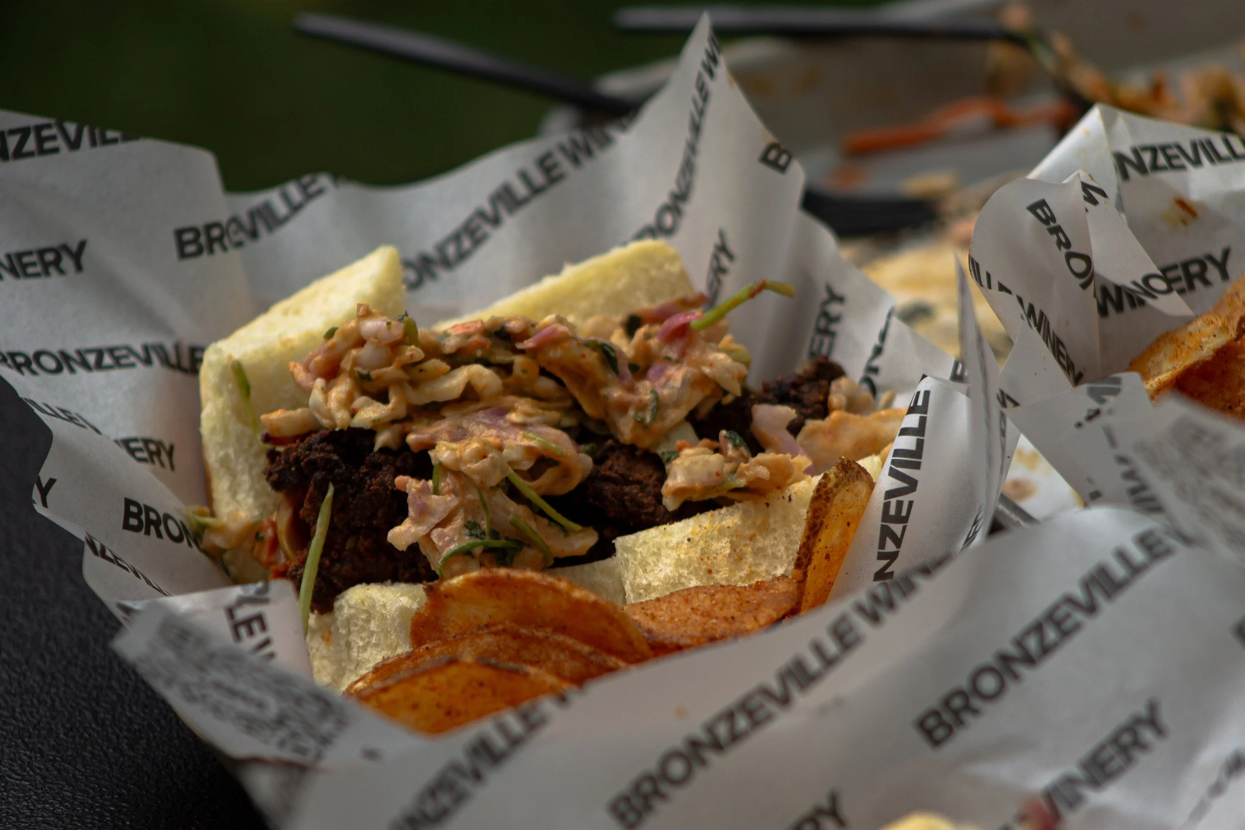 A close-up of a taco with shredded meat and shredded cabbage, wrapped in branded paper from Bronzed Valley. Some potato chips are visible nearby.