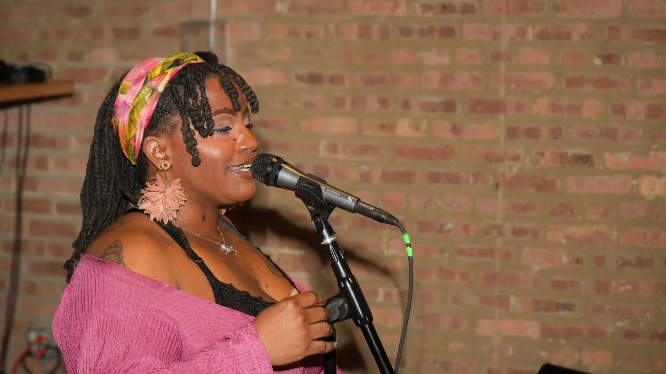 A woman with dreadlocks wearing a pink headscarf and pink earrings singing into a microphone against a brick wall background.