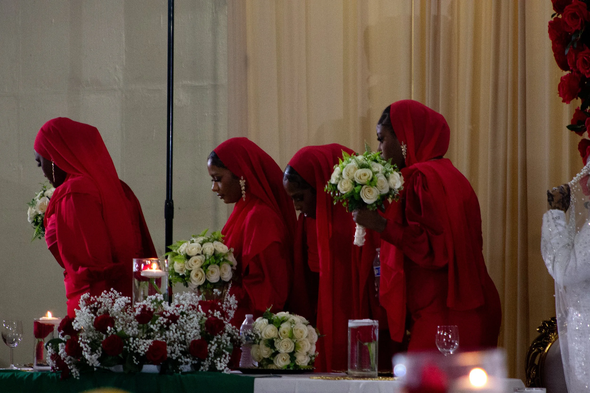 Four women dressed in red headscarves and dresses, standing behind a table with white and red floral arrangements and lit candles, in a formal indoor setting.