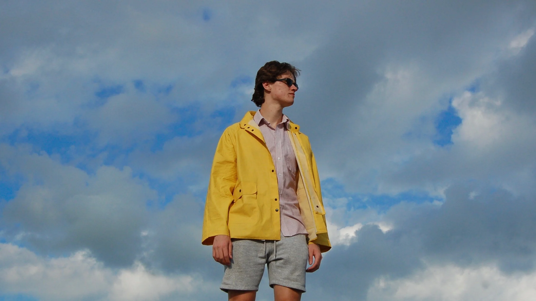 A young man wearing sunglasses, a yellow jacket, a striped shirt, and gray shorts standing outdoors against a cloudy sky background.