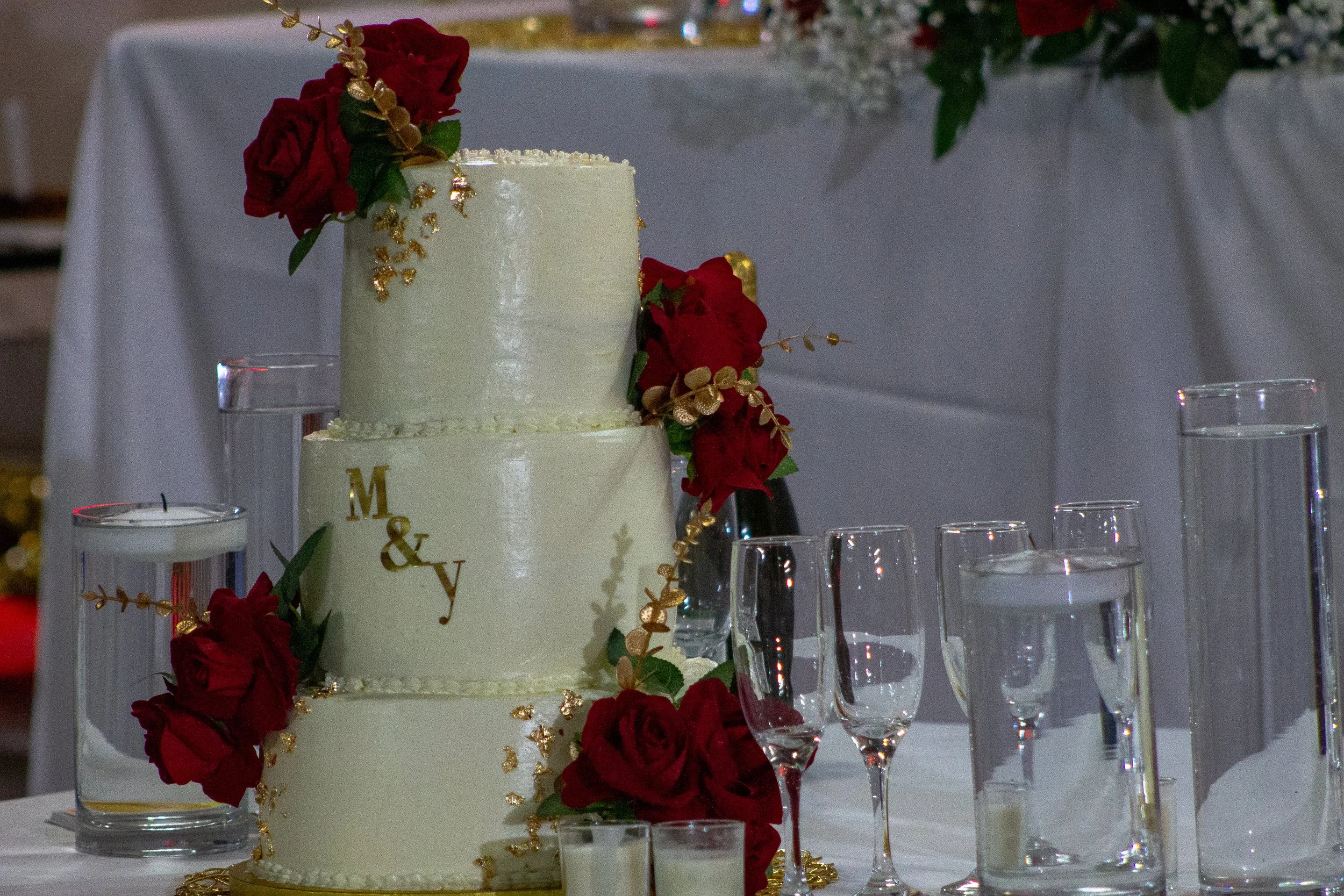 A two-tier white wedding cake decorated with red roses, gold accents, and greenery, placed on a table with champagne flutes and candles.