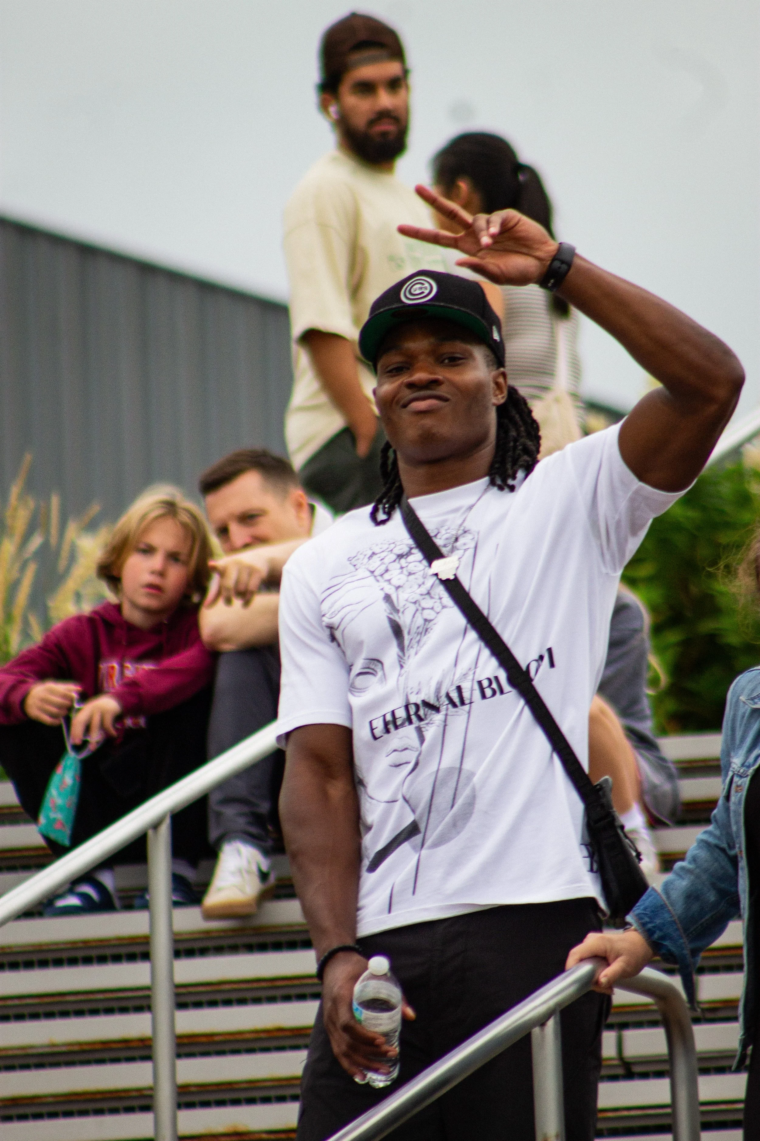 A young man with long dreadlocks wearing a white T-shirt and a black cap making a peace sign with his hand while standing on stairs, holding a water bottle. In the background, there are several other people, including a man with a beard and a baseball cap, a woman with a ponytail, and a young girl with short hair, sitting on the stairs.