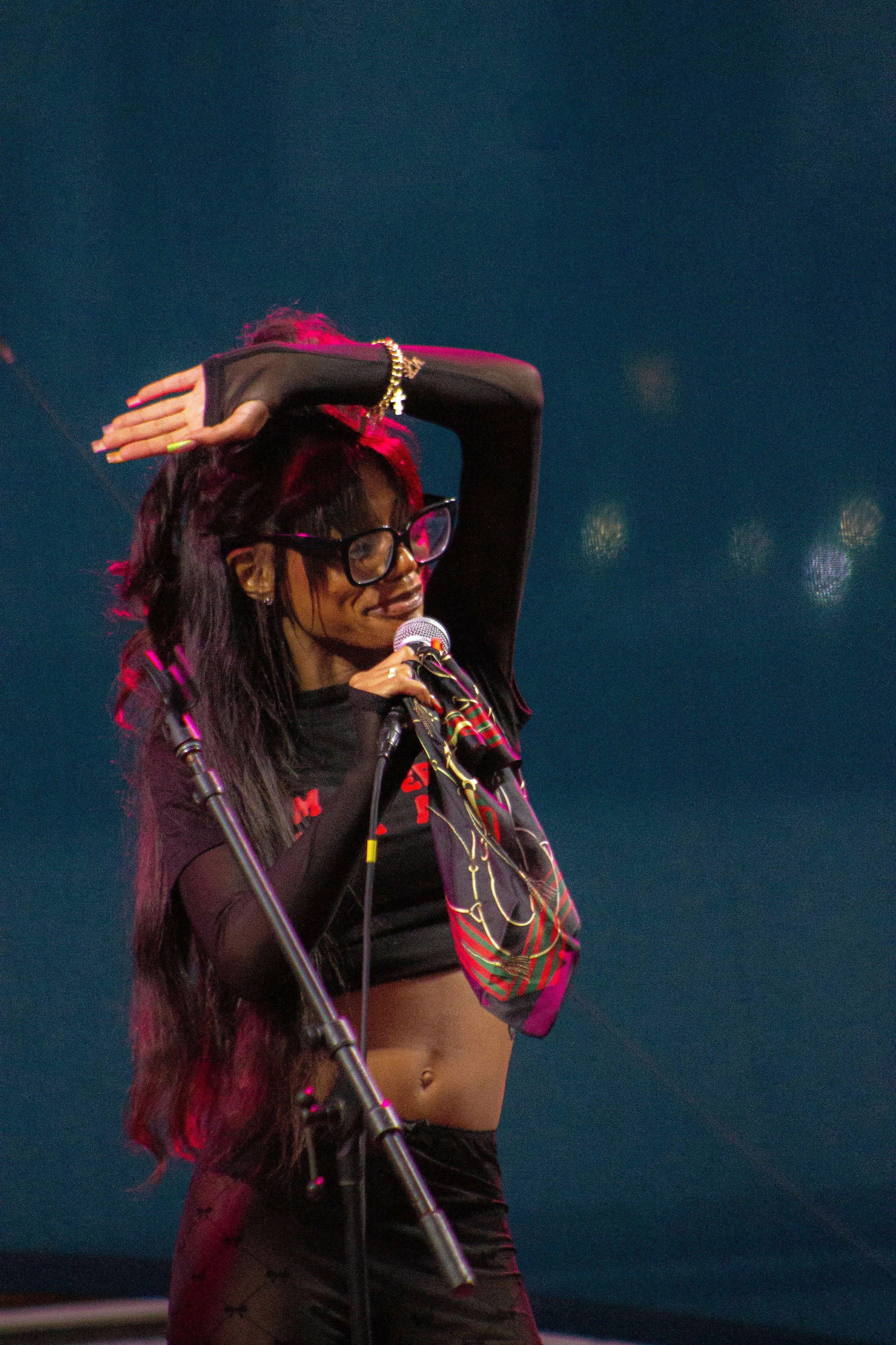 A woman with long dark hair and glasses, wearing a black crop top and patterned scarf, singing into a microphone with her left arm raised over her head. The background is dark with some blurry lights.
