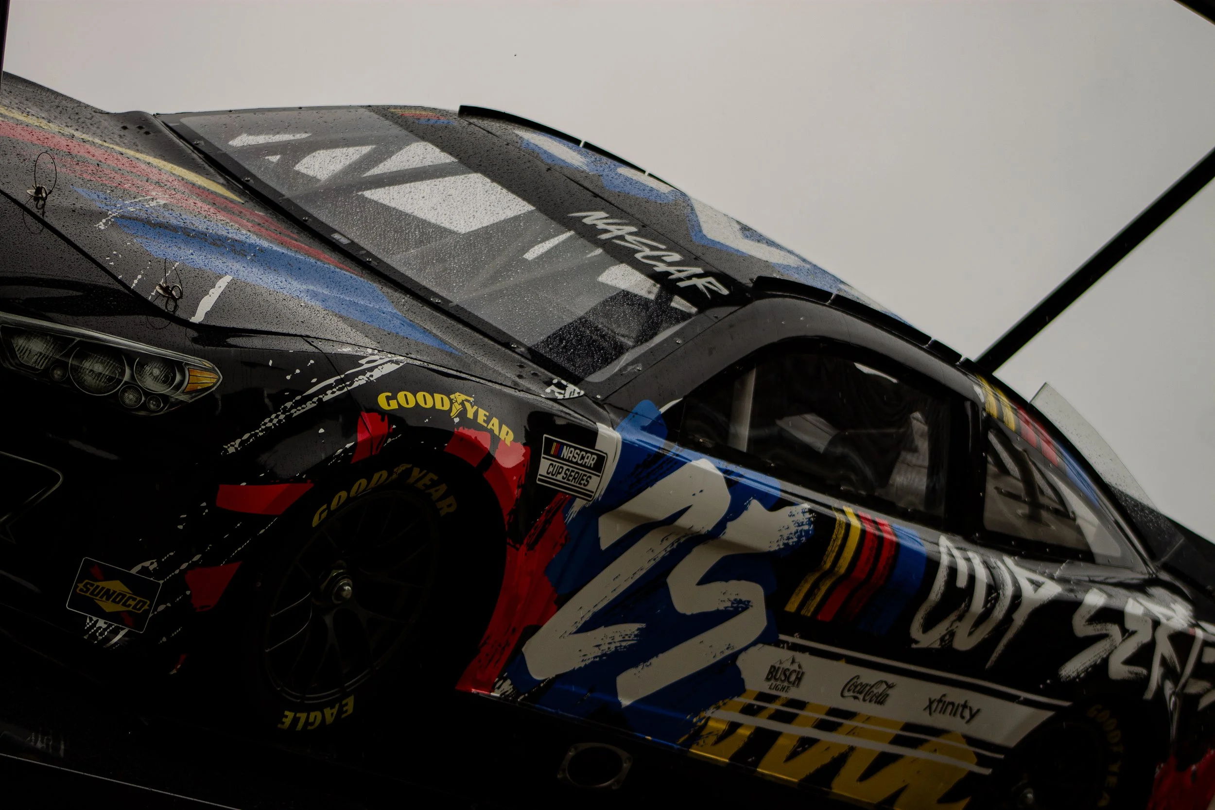 Close-up of a race car with sponsorship logos including Goodyear, Coca-Cola, Busch Light, and Xfinity, showing the windshield and front portion, with raindrops on the glass.