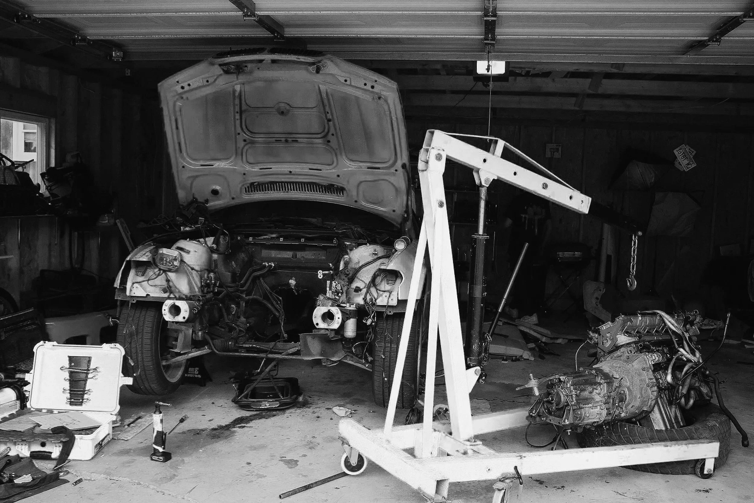 A garage workspace with a car being worked on, car parts and tools scattered around, and a hydraulic lift holding the car's front end