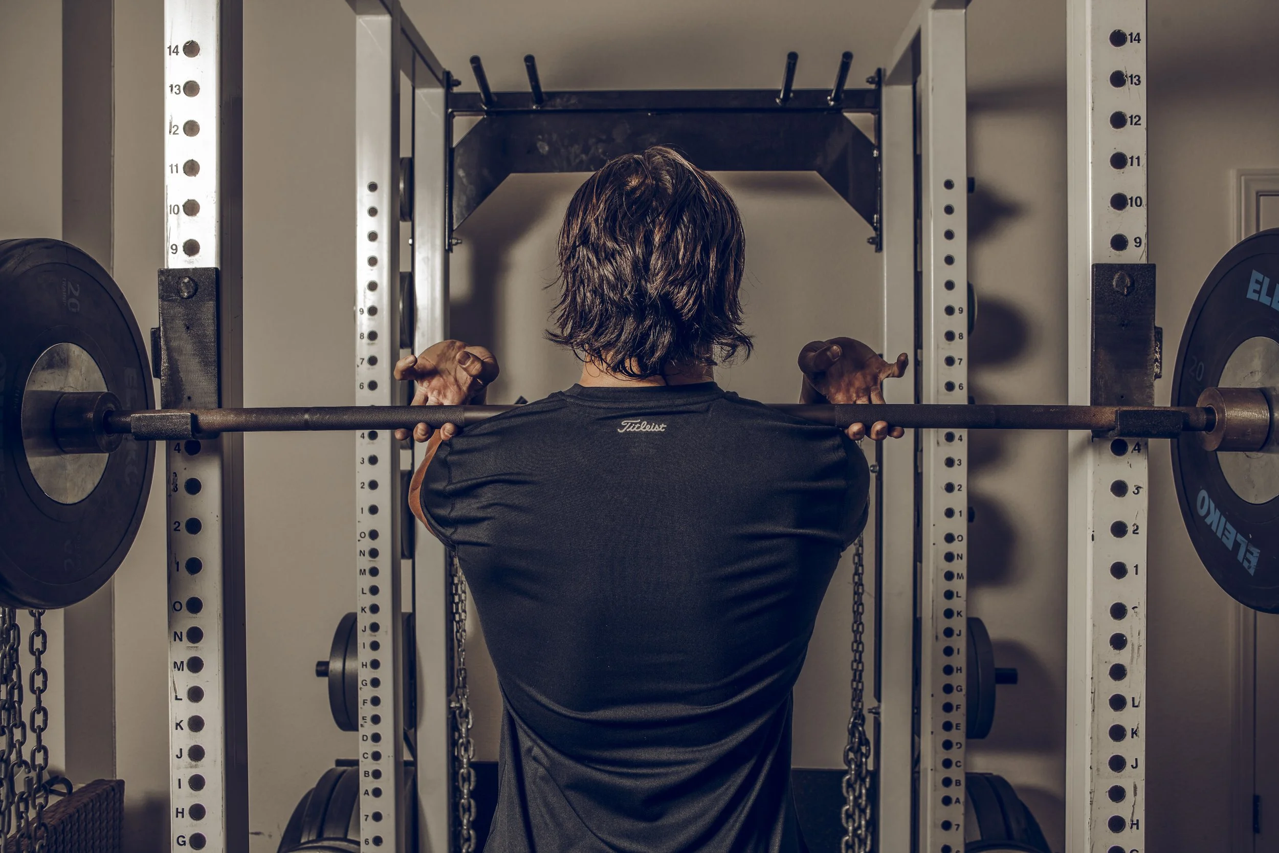 Person with wavy hair performing a squat exercise with a barbell on their shoulders in a home gym, surrounded by weight plates and equipment.