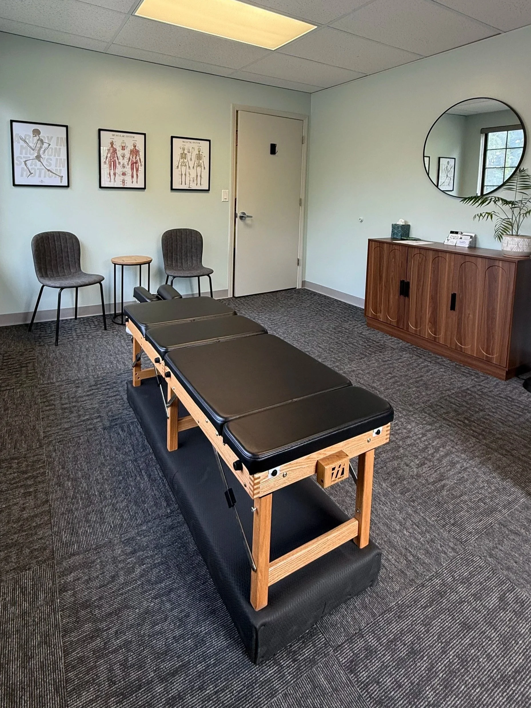 A therapy or chiropractic room with a massage table, two chairs, a small table, a cabinet, a mirror, and framed medical diagrams on the wall.