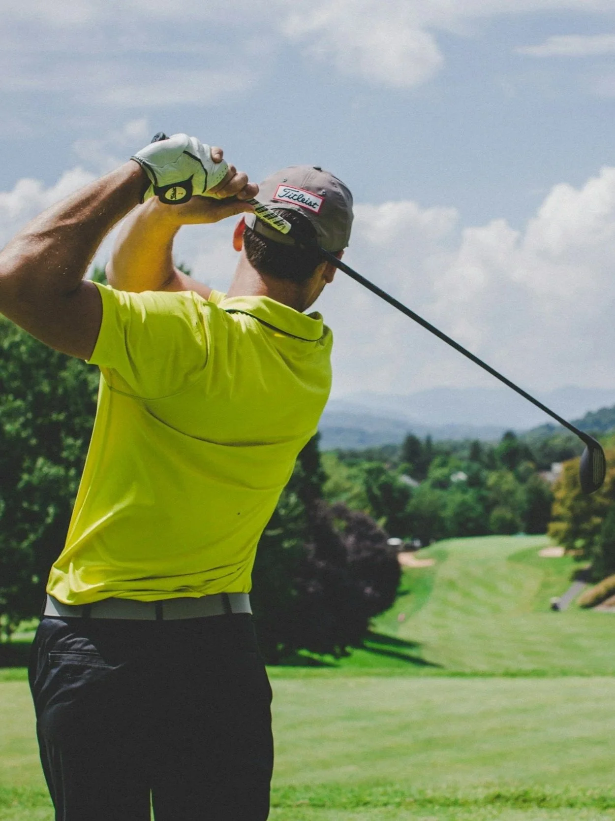 Man playing golf on a sunny day, swinging a golf club on a golf course with green grass and trees in the background.