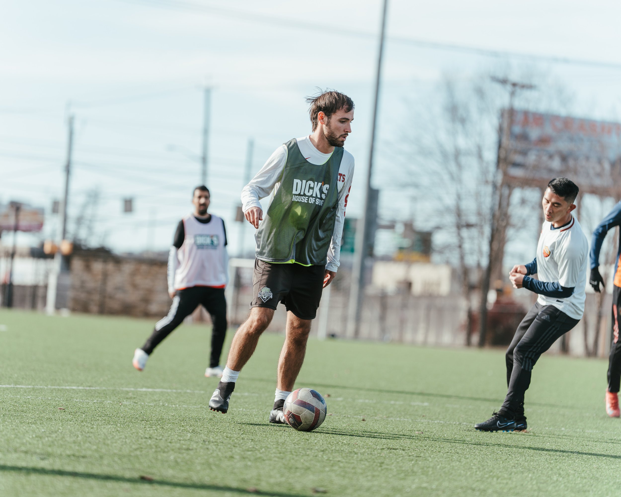 A man in a gray sports vest is on a soccer field, preparing to kick a soccer ball, with other players around him during daytime.