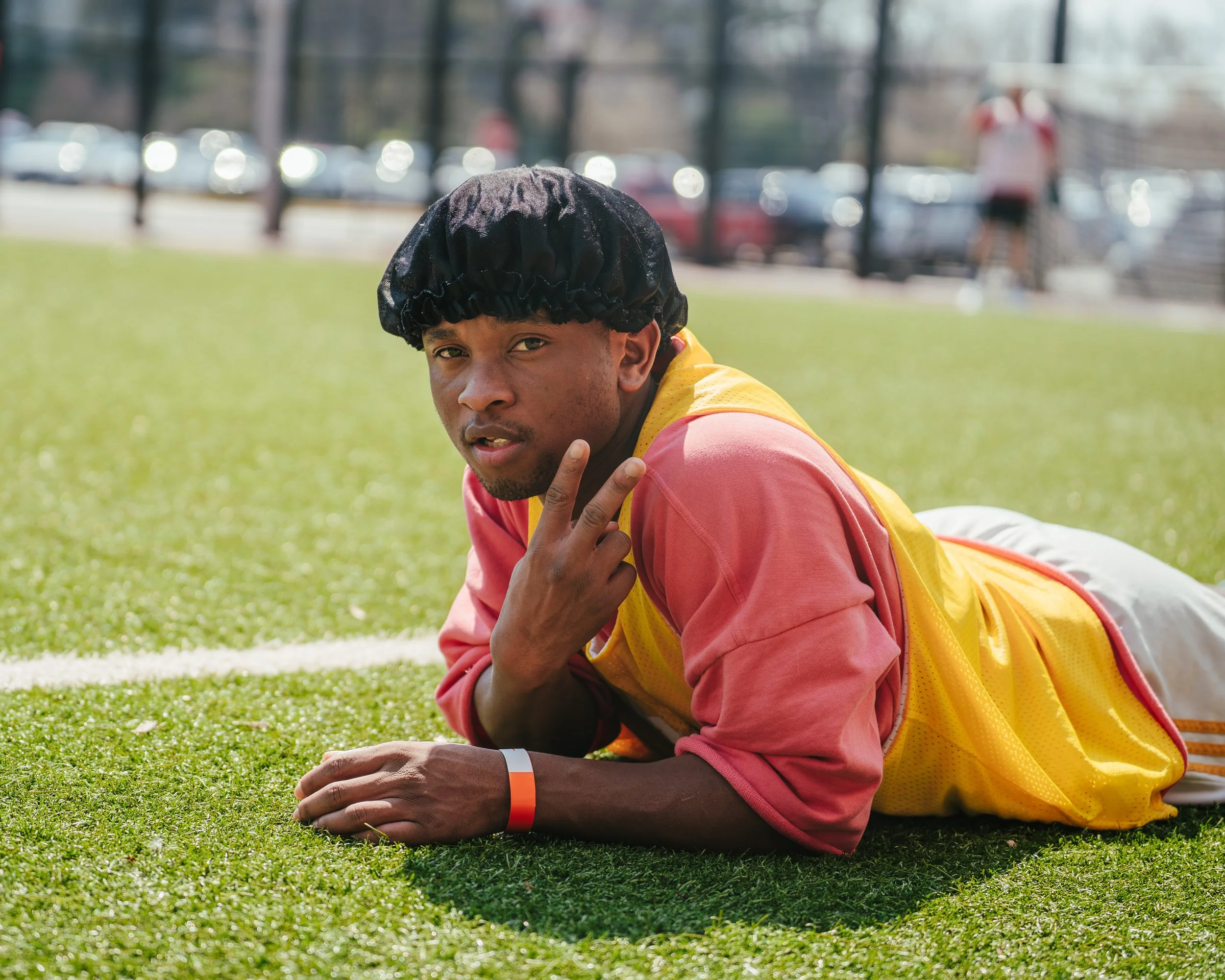A young man lying on the grass at the sports field, wearing a hair net, pink long-sleeve shirt, yellow sports vest, and wearing a red and white wristband, making a peace sign gesture.