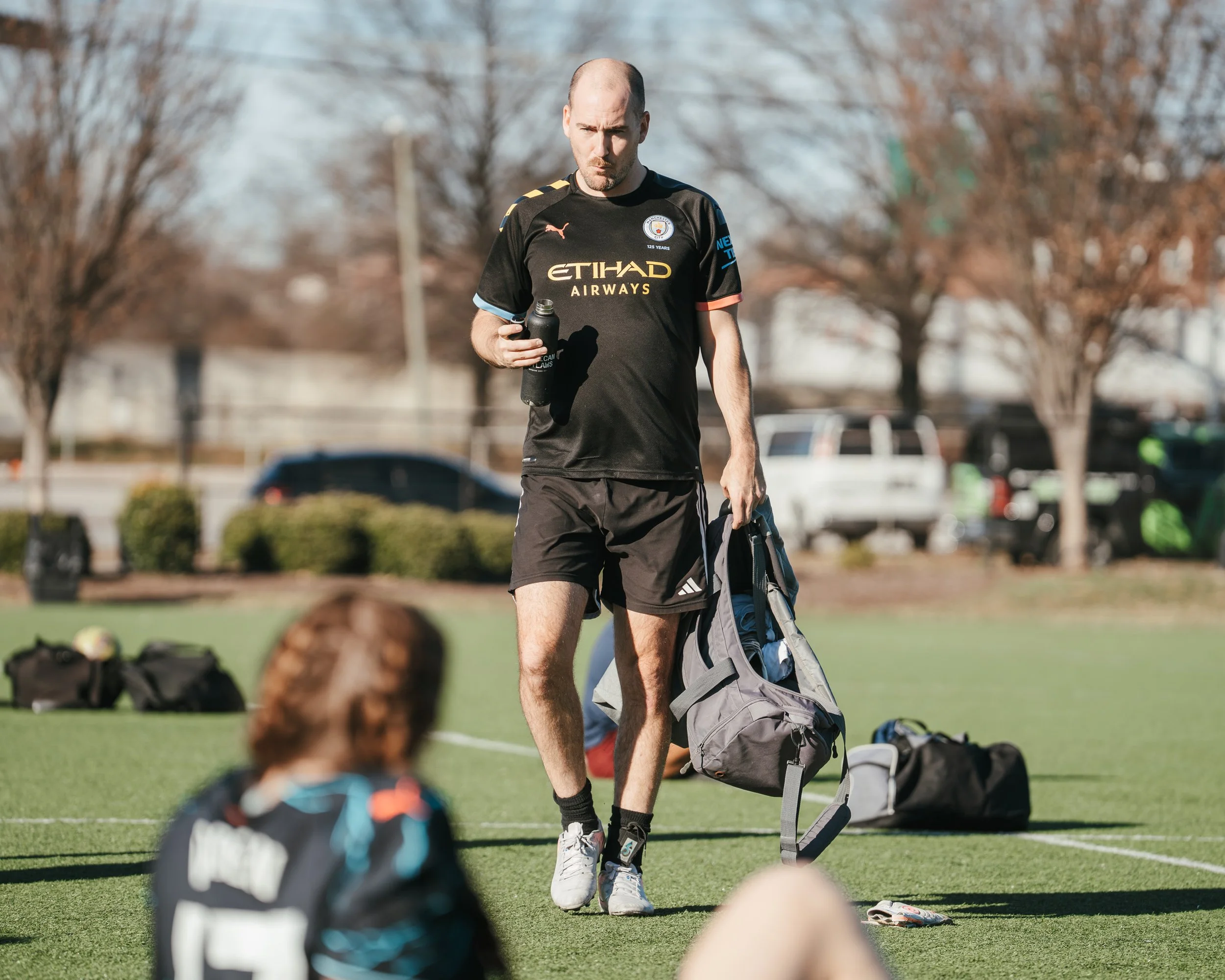 A man wearing a black Manchester City football jersey holding a sports drink bottle and carrying a sports bag, walking on a green field with children and sports equipment nearby, in an outdoor setting with trees in the background.