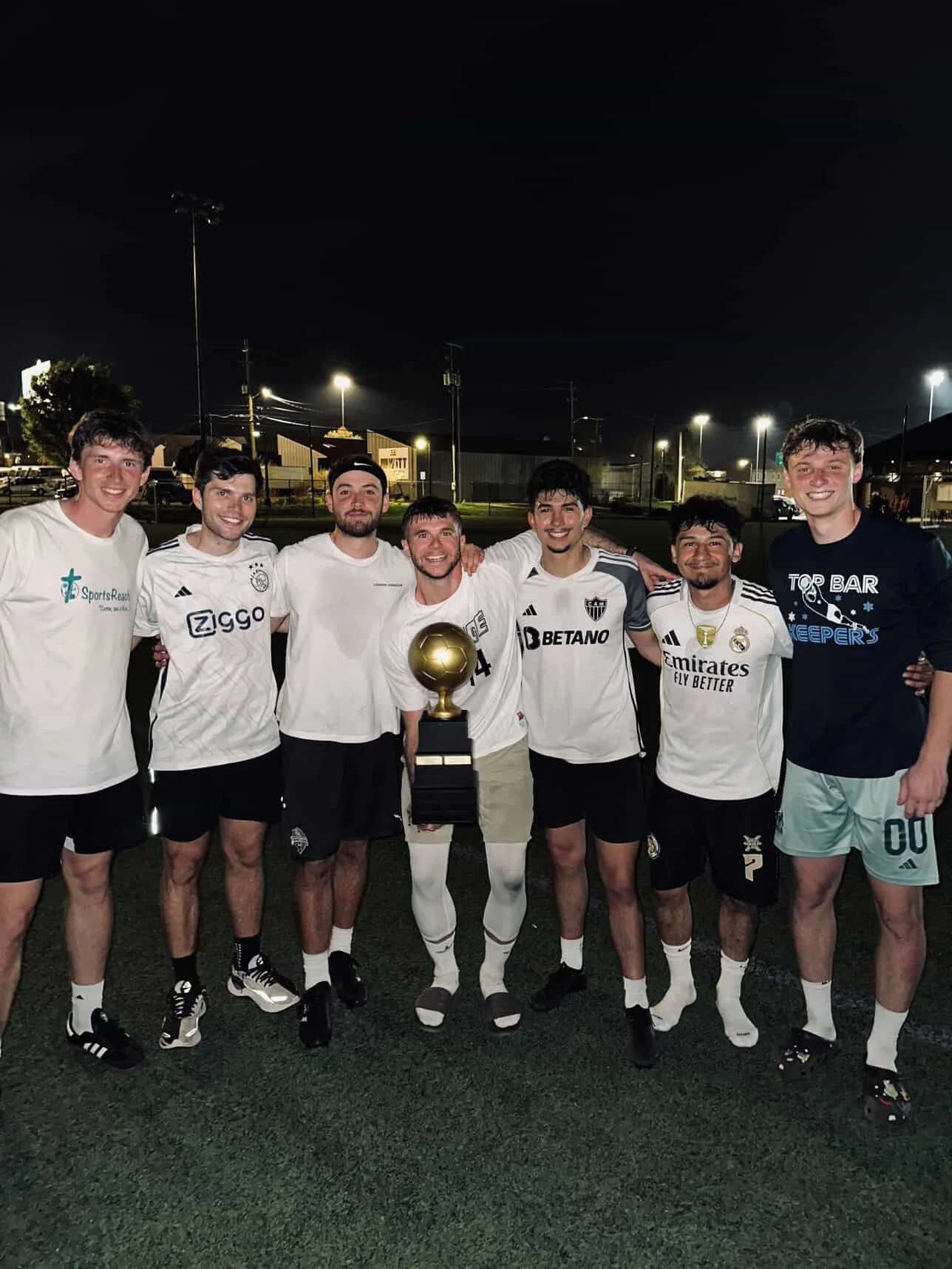 Group of seven young men standing outdoors at night, celebrating and smiling, with one holding a trophy. They are wearing soccer uniforms and casual clothing, standing on turf field.
