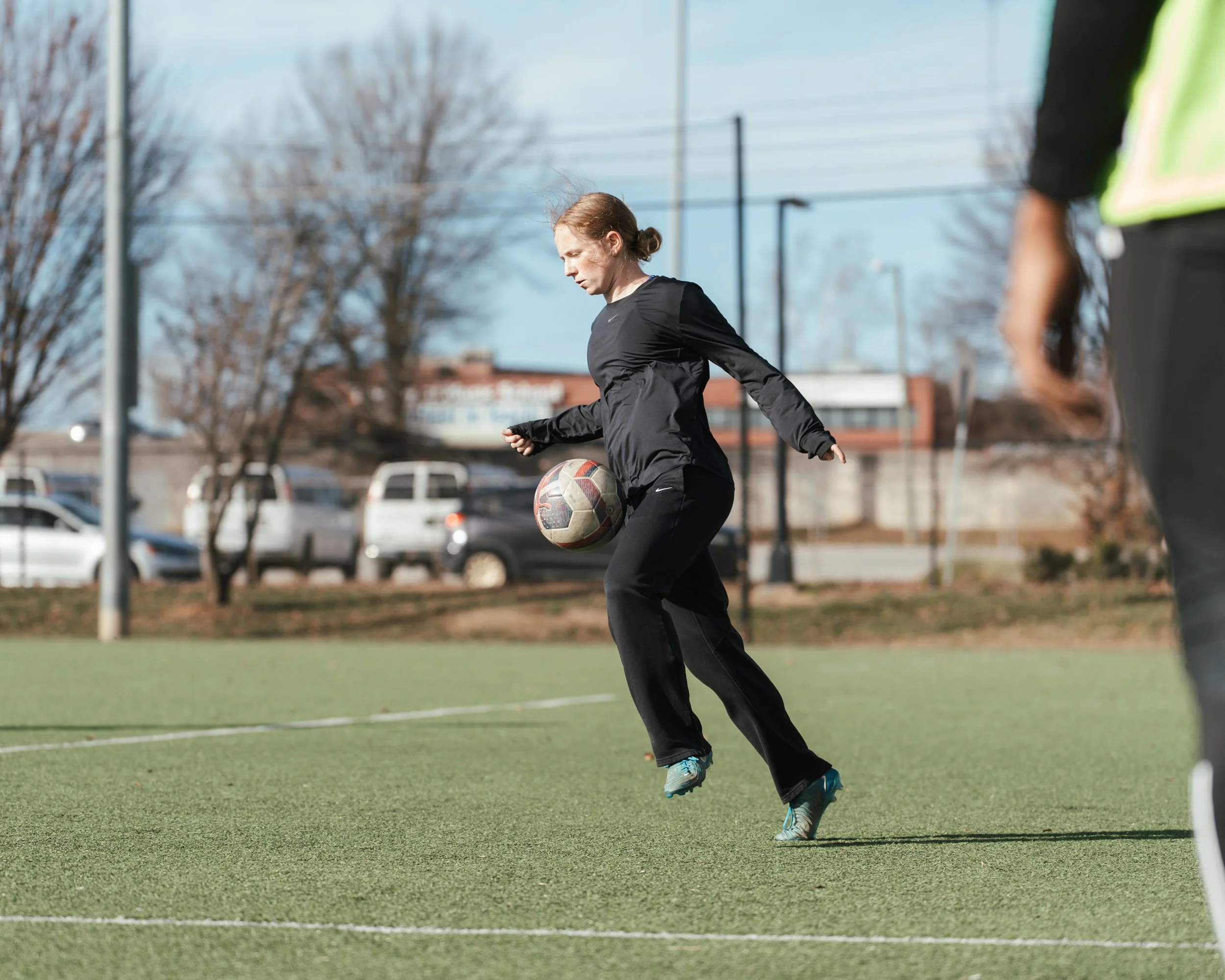 A woman in black athletic clothing dribbling a soccer ball on a green field with a goal in the background, outside on a clear day.