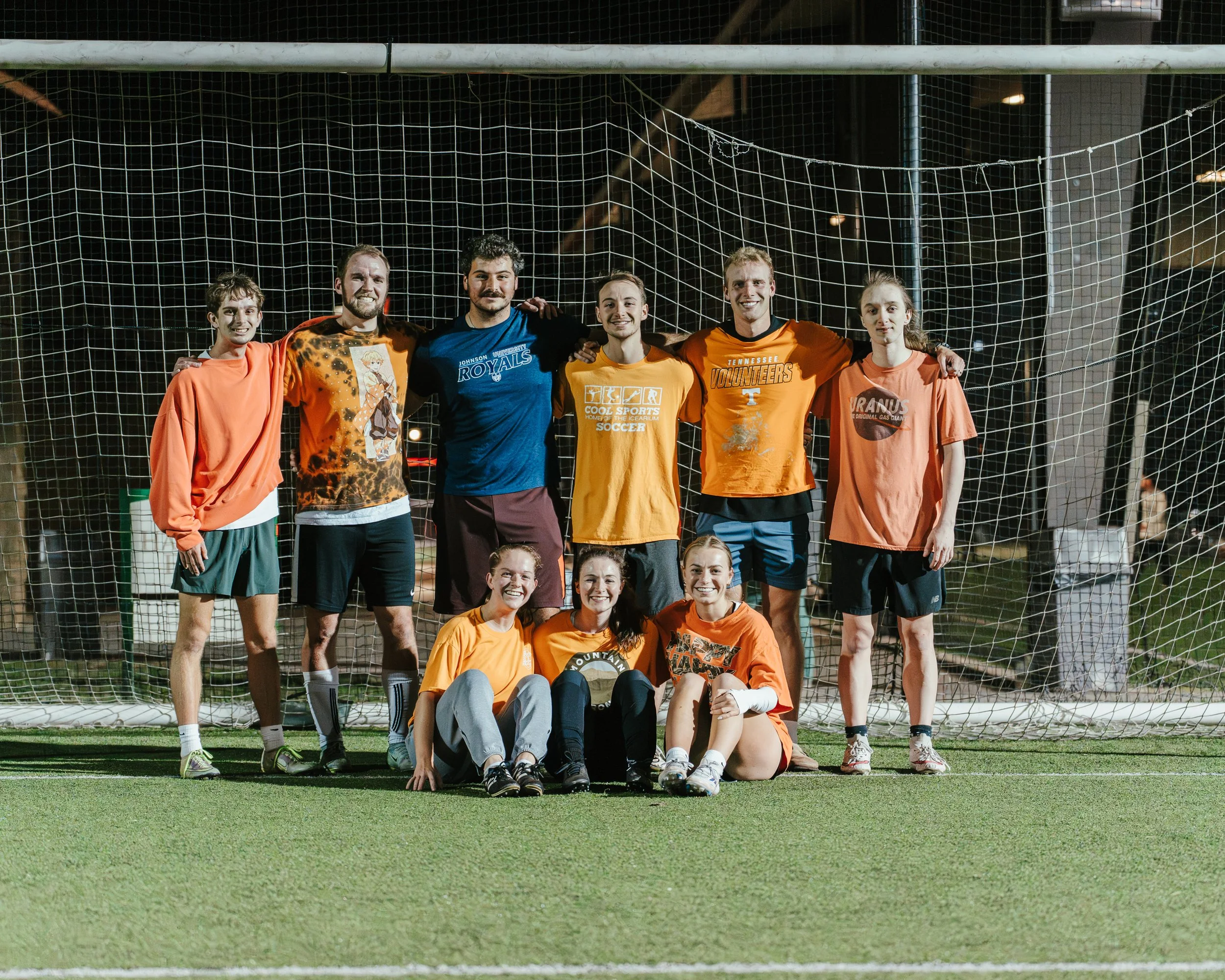 Group of young adults and teenagers posing in front of a soccer goal on a field at night, celebrating after a game.