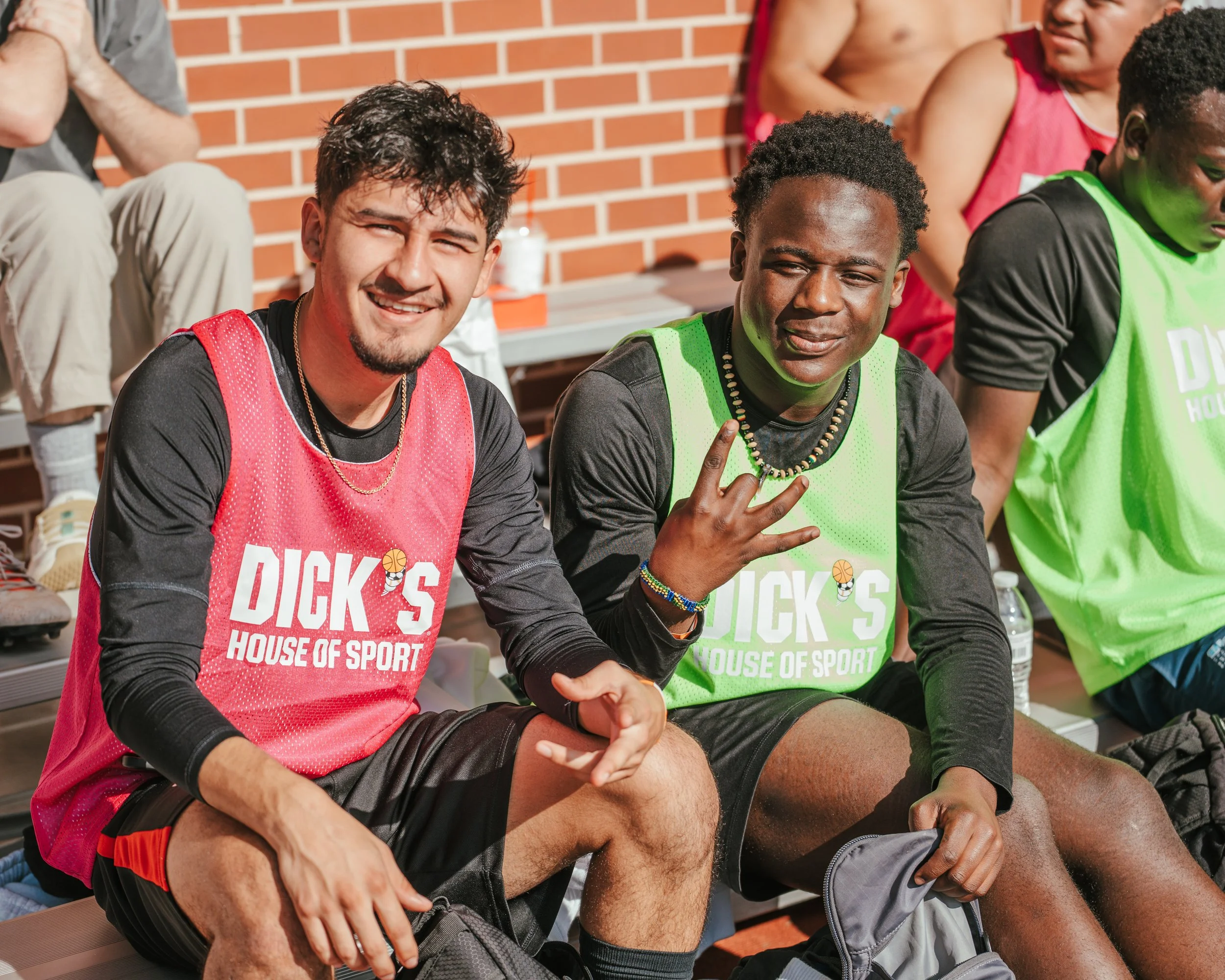Group of young people sitting on a bench during a sports event, wearing colorful sports jerseys with 'DICKS House of Sport' printed on them, with a brick wall in the background.