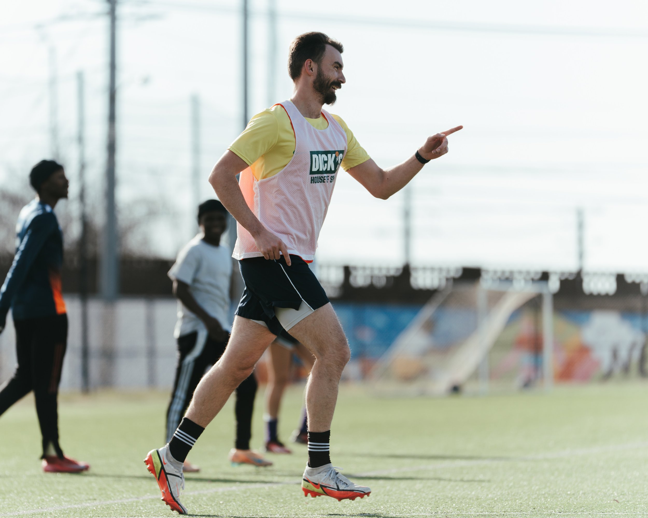 A group of people playing soccer outdoors, with one man in the foreground wearing a yellow shirt, a white and pink training bib, black shorts, and gray and red shoes, running and pointing during the game.