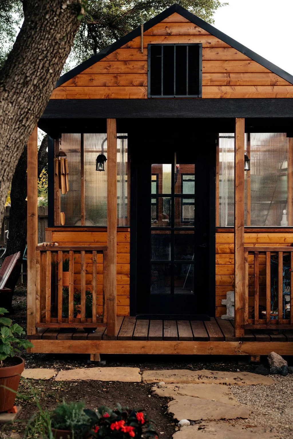 A small wooden cabin with a black door, wooden railing, and a small porch, surrounded by trees and garden.