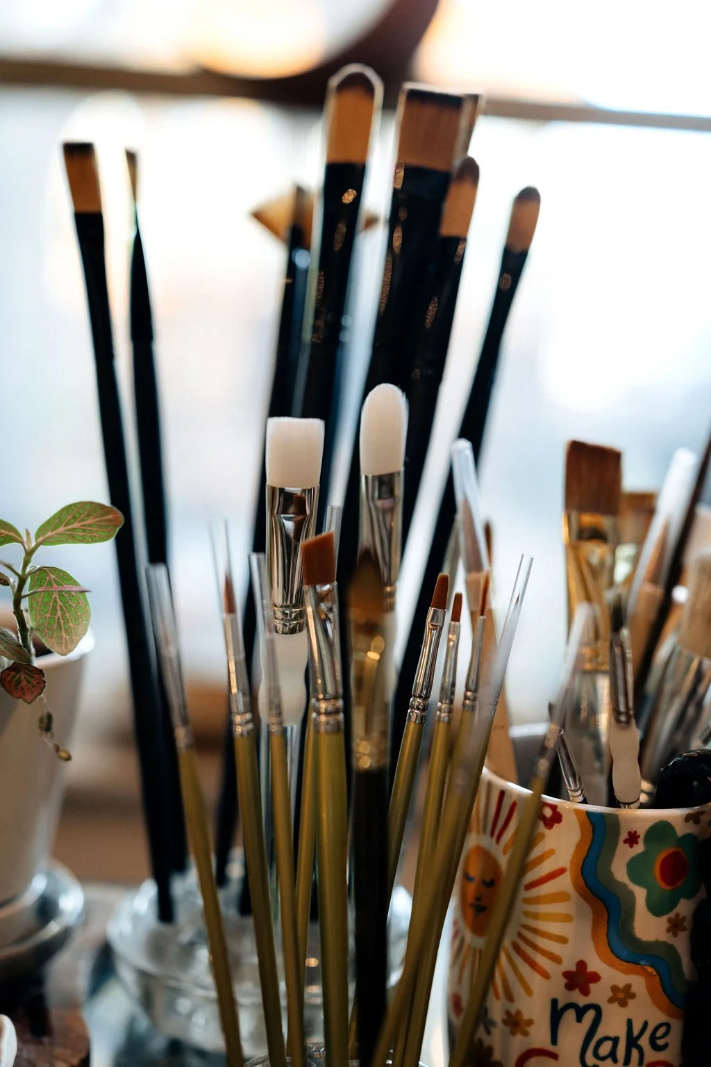 Close-up of various paintbrushes in a decorative mug and a jar, with a potted plant visible on the side.