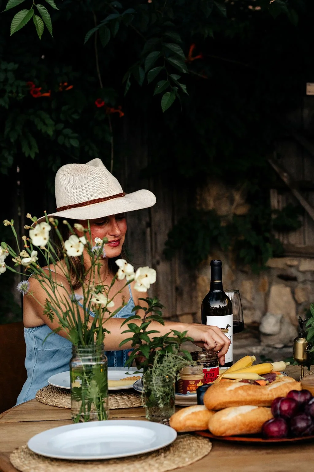 A woman wearing a wide-brimmed hat and a blue dress sits at an outdoor table with bread, cheese, grapes, wine, and flowers, surrounded by greenery.