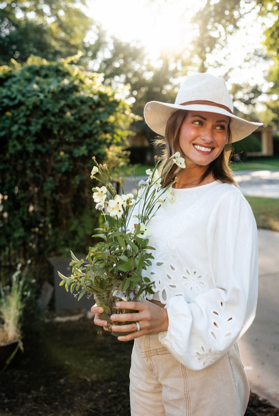 A woman smiling, wearing a white sun hat and a white embroidered blouse, holding a glass vase with white flowers outdoors during daylight.