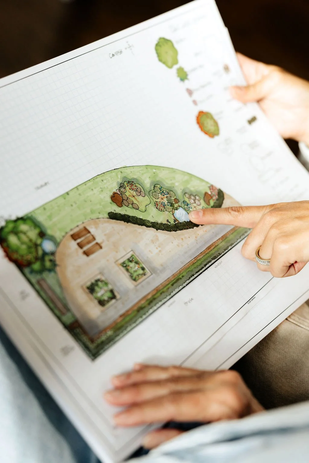 A person pointing at an illustrated landscape plan in a book, showing a green garden with trees, bushes, a house, and artwork on the walls.