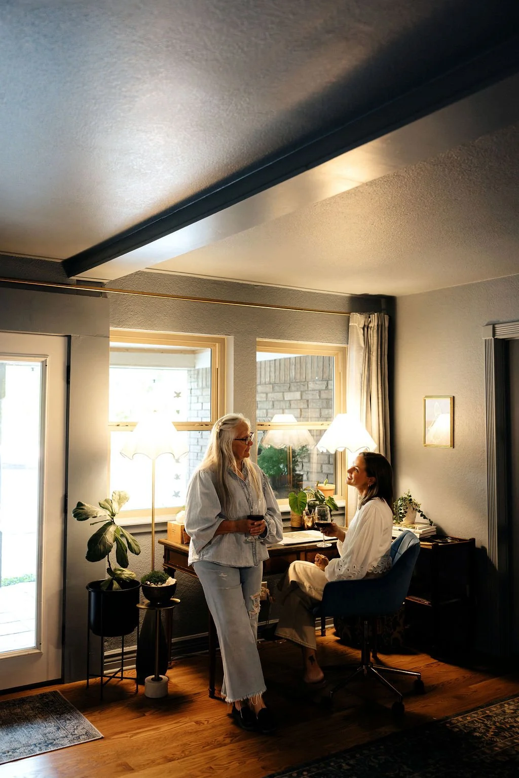 Two women in a cozy living room having a conversation, one standing and holding a glass of wine, the other sitting in a blue chair with a glass of wine, surrounded by houseplants and warm lighting from lamps.