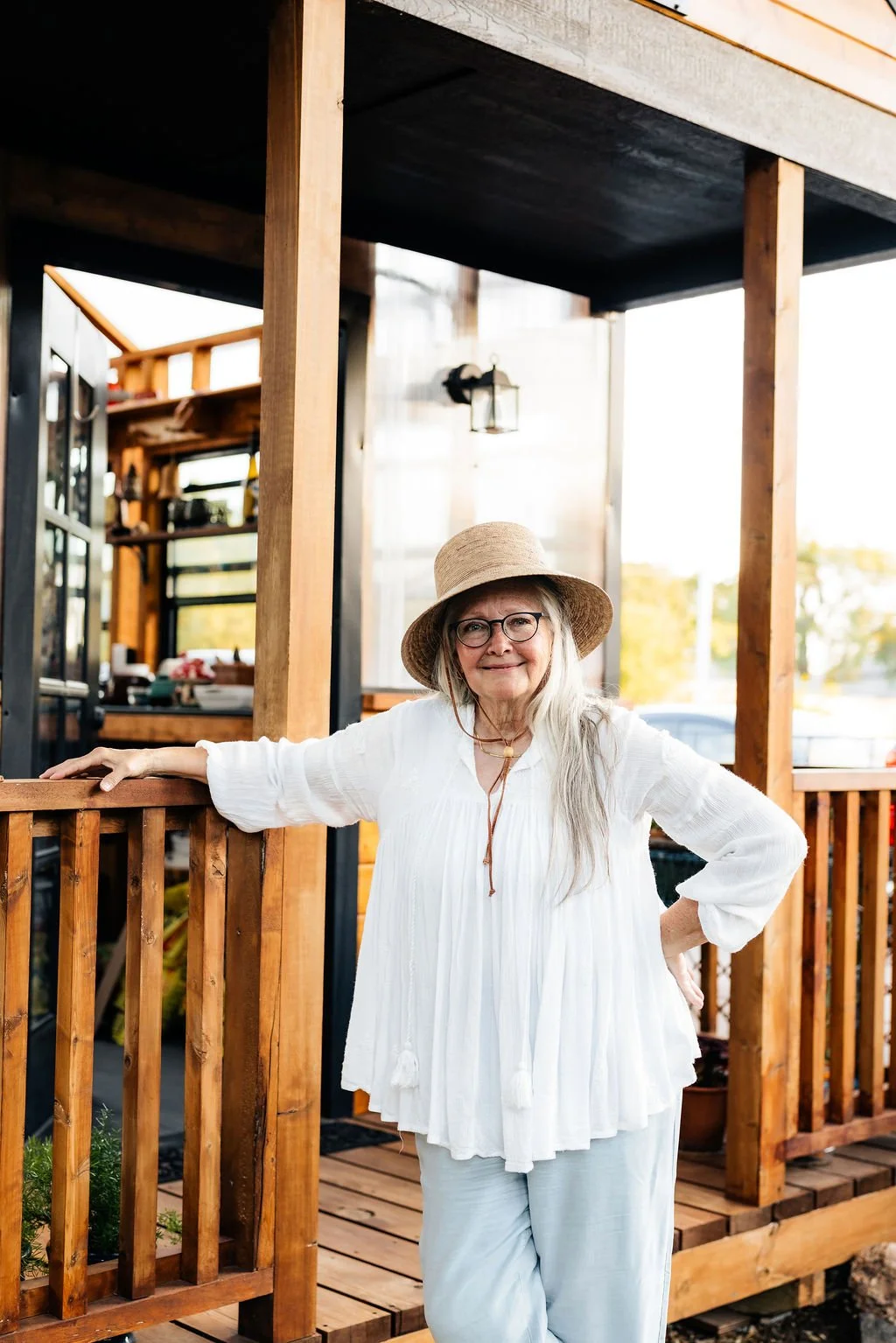 An elderly woman with long gray hair, glasses, and a wide-brimmed hat standing on a wooden porch with a relaxed pose and smiling. She is wearing a white blouse and light-colored pants.