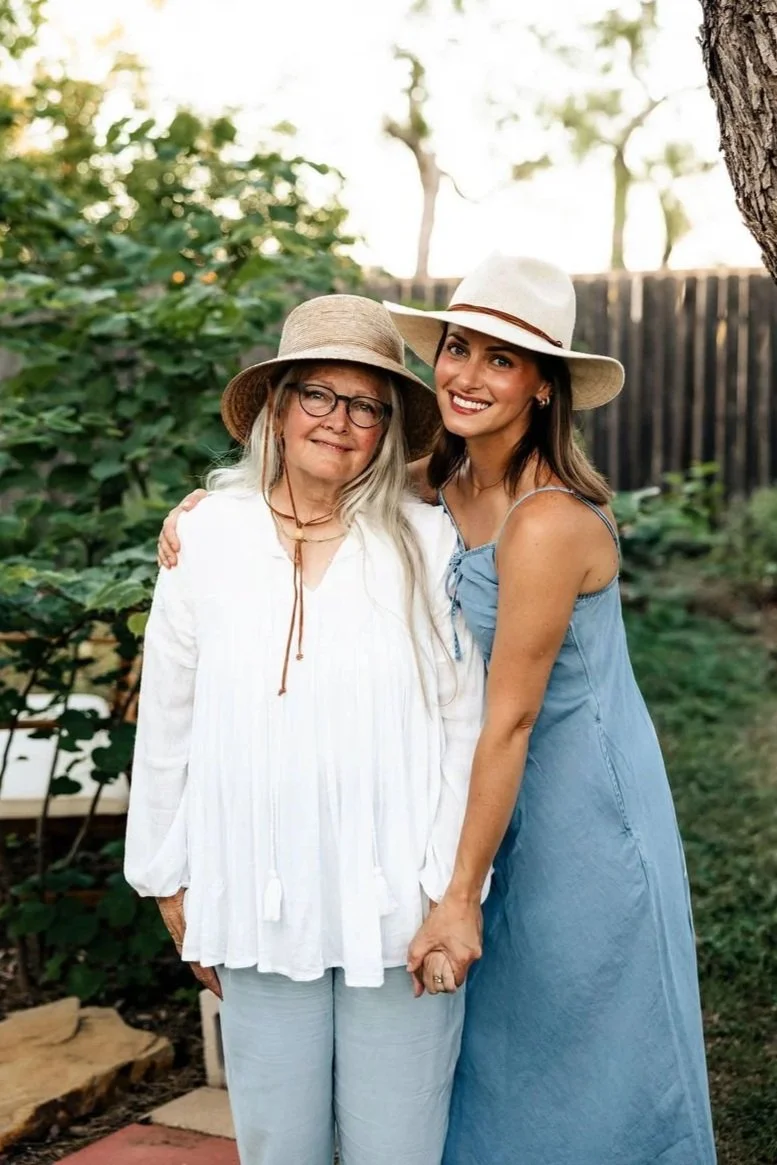 Two women standing outdoors, smiling and holding hands, wearing sun hats, with a tree and wooden fence in the background.