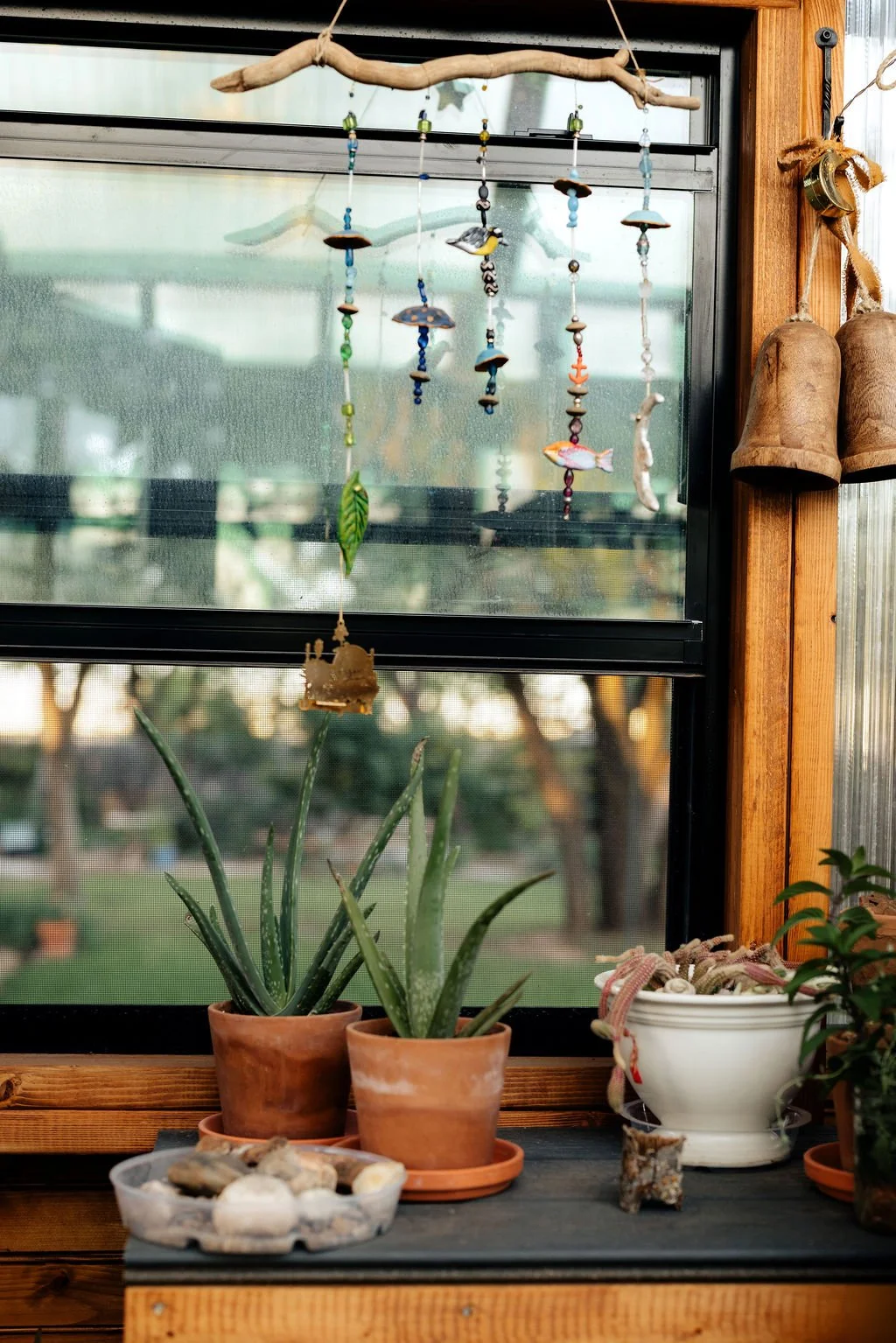 Decorative garden scene viewed through a window with hanging ceramic wind chimes and potted plants on a windowsill.