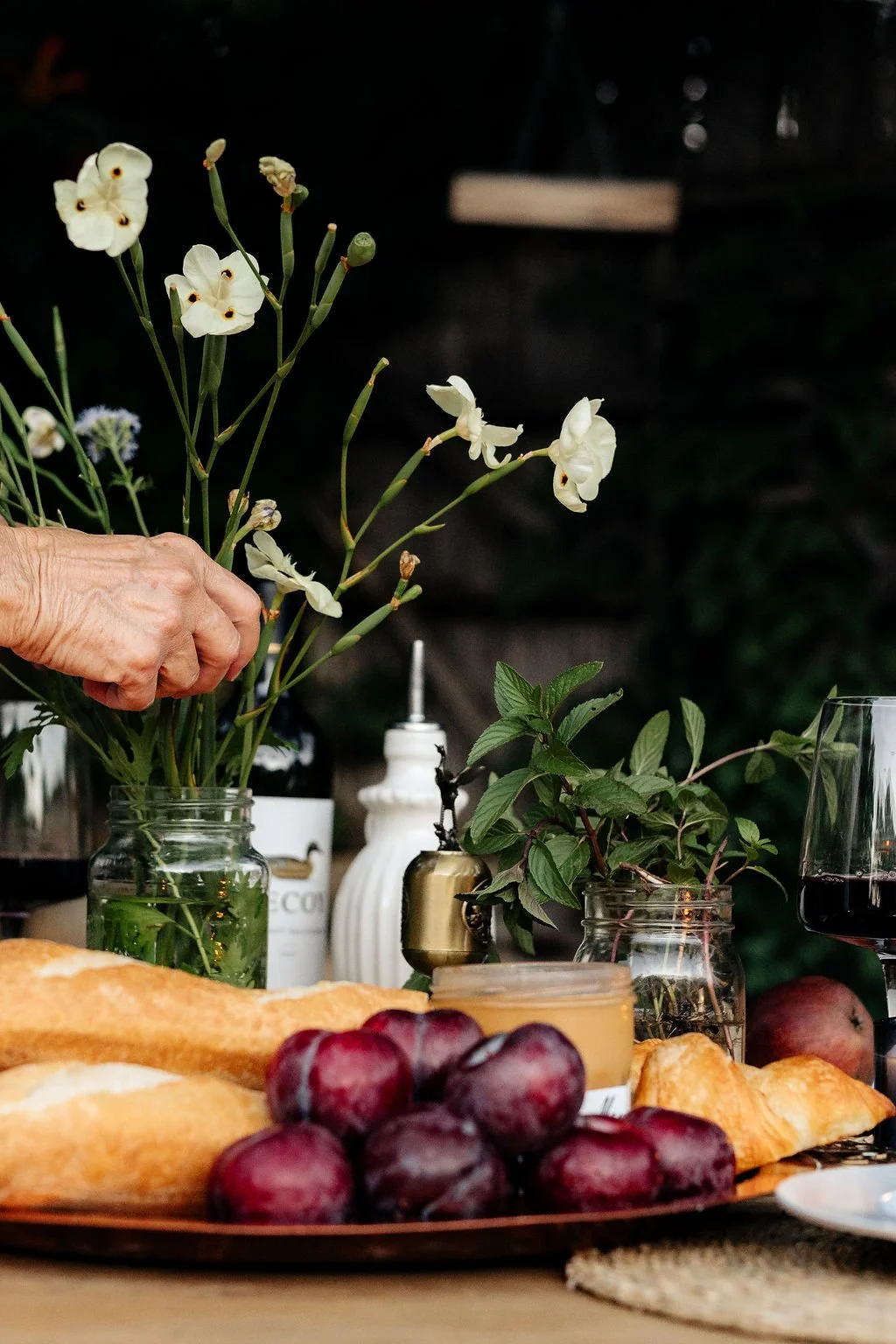 Close-up of a table with fresh flowers, grapes, croissants, jam, red wine, and greenery, with a hand arranging flowers in a jar.