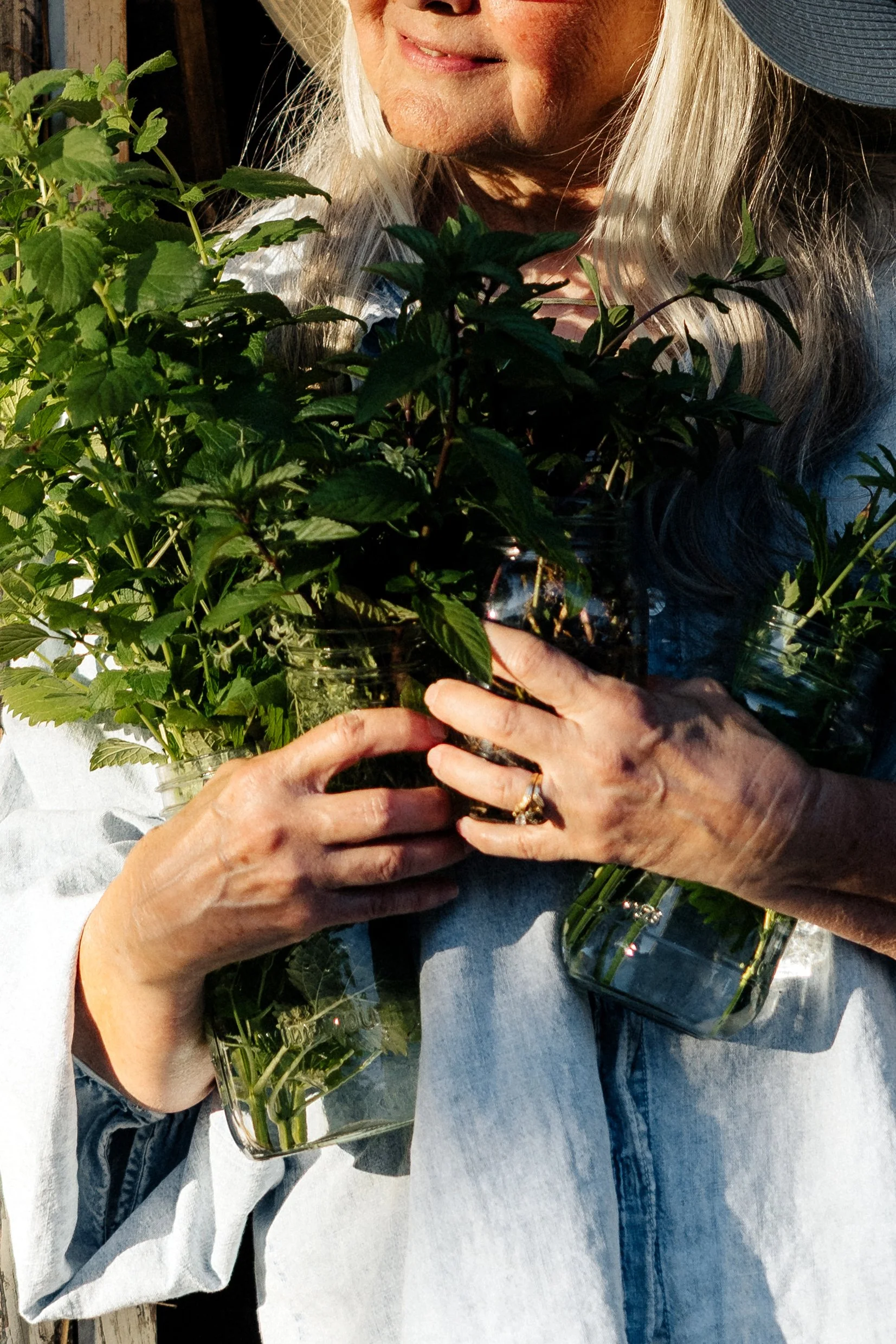 Close-up of an elderly woman with long gray hair holding a small potted plant and a glass jar with green plant cuttings, wearing a light-colored shirt and blue jeans.