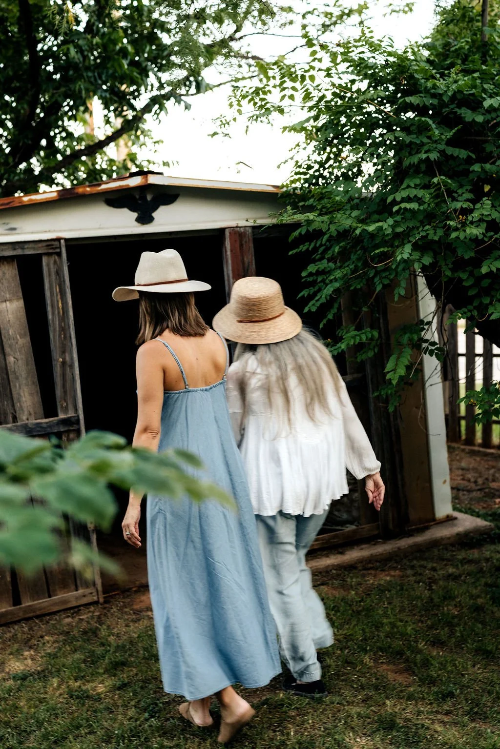 Two women wearing wide-brimmed hats walking towards a rustic shed surrounded by greenery.